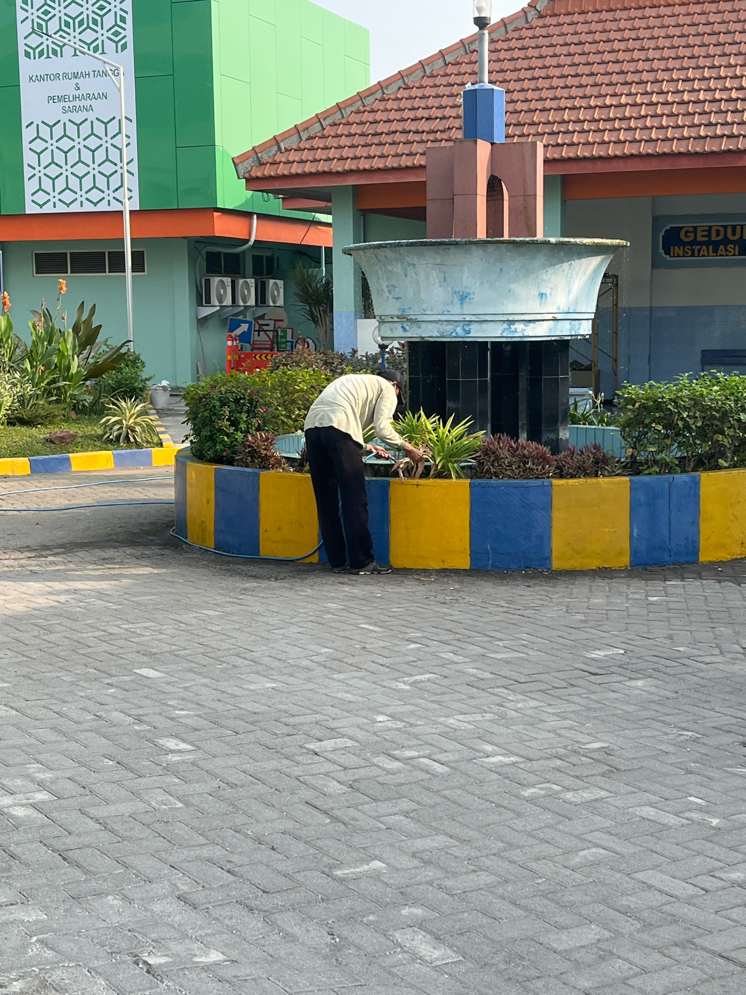 The image depicts a man in a light-colored long-sleeved shirt and dark pants tending to plants in a circular planter bed. The planter bed is bordered by alternating yellow and blue painted concrete blocks. A blue hose is visible on the ground, leading towards the planter. In the background, a building with a green facade and orange trim is visible, with text that reads "KANTOR RUMAH TANGG & PEMELIHARAAN SARANA." To the right, a large, light blue fountain basin with a tiered brown and blue structure on top is present. The ground is paved with grey interlocking bricks. The scene appears to be outdoors, possibly at an institution in Lamongan, Indonesia, given the Indonesian text on the building.