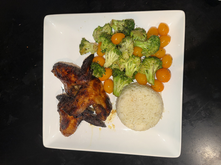 A white, rectangular plate containing a meal is positioned centrally on a dark, uniform surface, likely a countertop or table. On the upper left of the plate, four pieces of cooked chicken wings are arranged, exhibiting a dark reddish-brown, slightly charred appearance with some visible drippings. To the lower left, a compact, dome-shaped portion of white rice is present. Occupying the right side of the plate is a mixed serving of green broccoli florets and numerous small, round, orange-yellow cherry tomatoes, seasoned with what appears to be dried herbs or spices. No individuals are depicted, and no actions or interactions are occurring. The lighting is uniform, suggesting an indoor setting. The specific location within Los Angeles, United States of America, cannot be determined from the visual information provided.