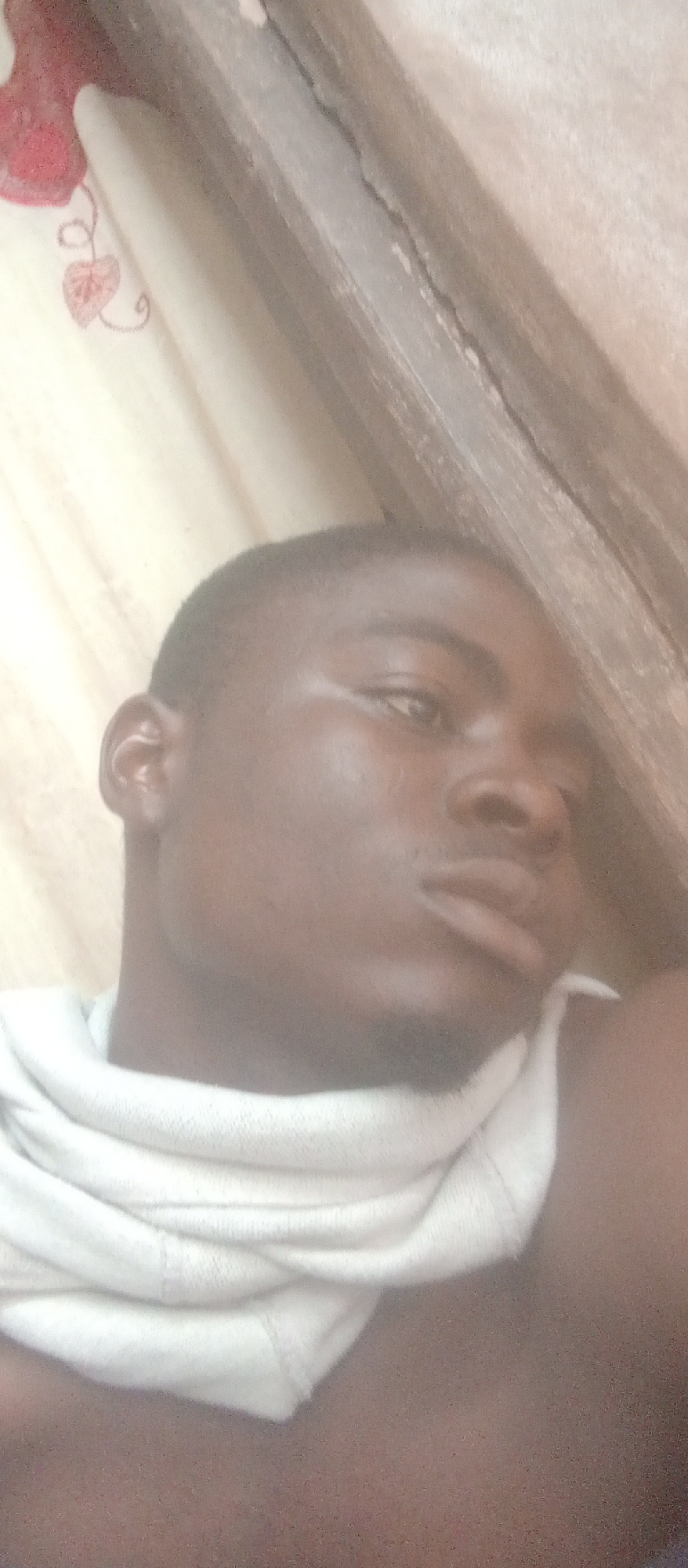 A close-up, slightly tilted, self-portrait of a young Black male lies on his side, leaning against a wooden structure. He is wearing a white garment that appears to be a t-shirt or sweater, pulled up around his neck. The background consists of a light-colored fabric with a subtle red floral pattern on the left side. The overall lighting is dim and appears to be from an artificial source. The image was taken in a domestic environment, likely a bedroom or living area in Ogidi, Nigeria.