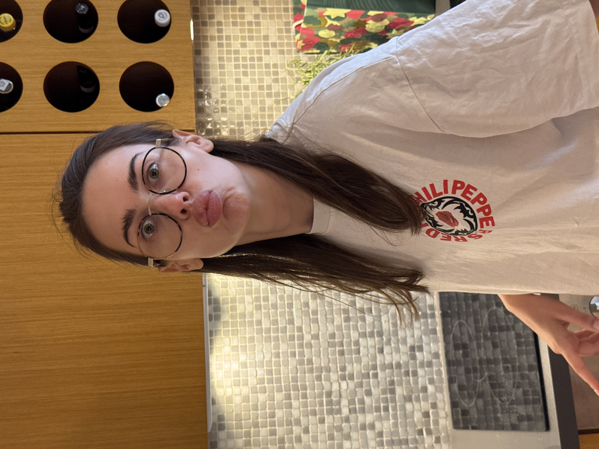 A woman with dark brown hair and round glasses is posing for a picture. She is wearing a white t-shirt with a "Red Chili Peppers" logo. She is making a duck face expression. Behind her is a wooden wine rack with several bottles, a mosaic tile backsplash, and a glimpse of a patterned blanket. The location is an indoor space with kitchen elements. The city and region are unknown.