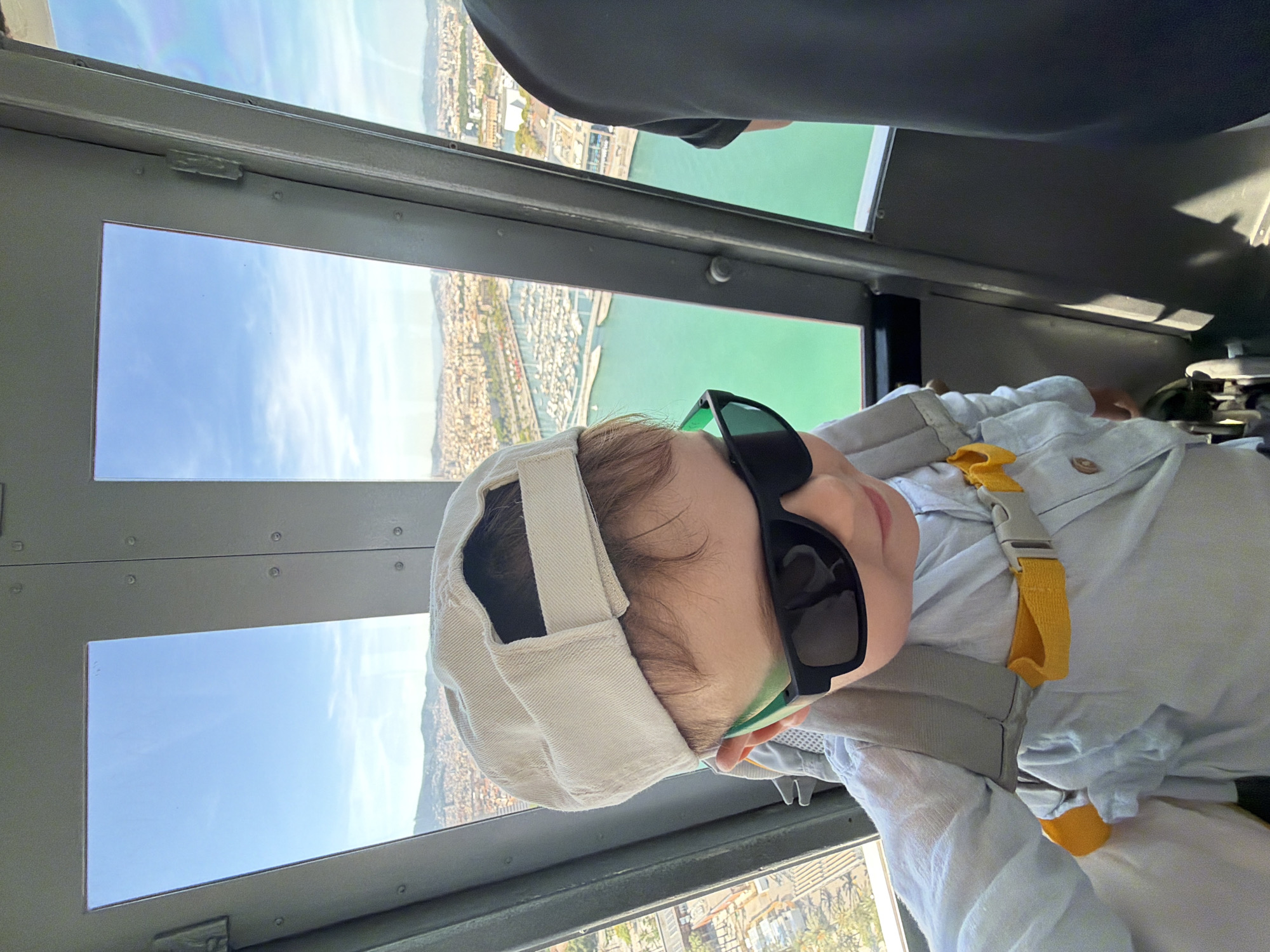 The image shows a young child wearing sunglasses and a baseball cap, looking out of a window. The child is in what appears to be a cabin or vehicle, with metal framing and large windows. Outside the window, a city skyline with buildings and a harbor with many boats is visible. The water in the harbor is a vibrant turquoise color. The child is wearing a light-colored shirt and a backpack. The setting is the Port Cable Car (Telefèric del Port) in Barcelona, Spain, offering panoramic views of the city and the coastline.