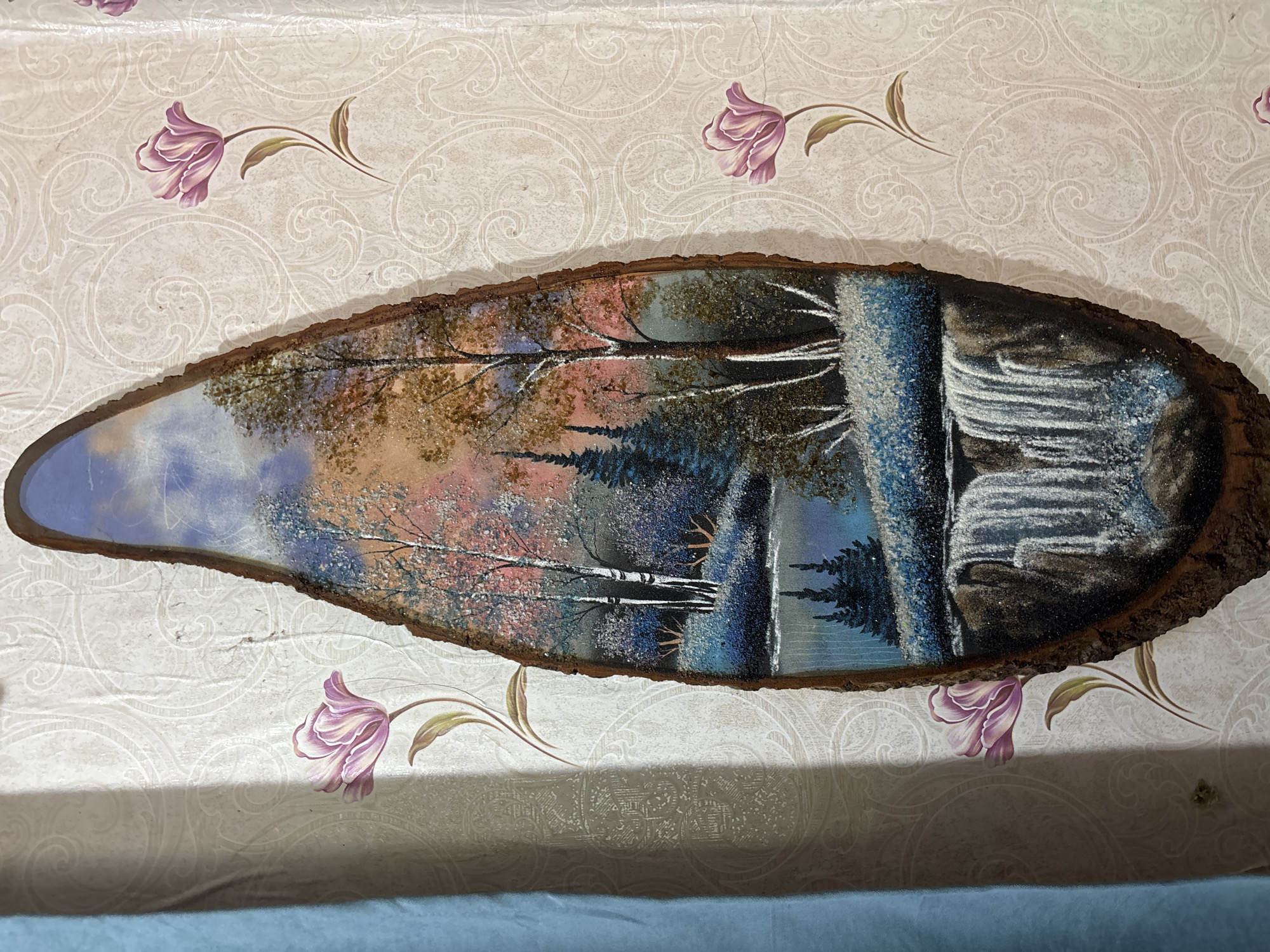 The image displays a painting on a piece of wood, depicting a natural landscape. The painting features a waterfall at the bottom, transitioning into a body of water with trees and a colorful sky above. The wood appears to be a slice from a tree trunk, with the bark still attached to the edges. The artwork is mounted on a wall with floral patterned wallpaper. A blue curtain is visible on the left side of the image, casting a shadow on the wallpaper. The painting itself is detailed and appears to incorporate glitter or small reflective particles, particularly in the snow-like areas and water. The colors are vibrant, with shades of blue, brown, white, and pink dominating the scene. There are no people or animals visible in the painting or in the surrounding environment. The image is taken from a slightly elevated angle, looking down at the artwork. The location in Zubov, Russia is not discernible from the visual information provided in the image.