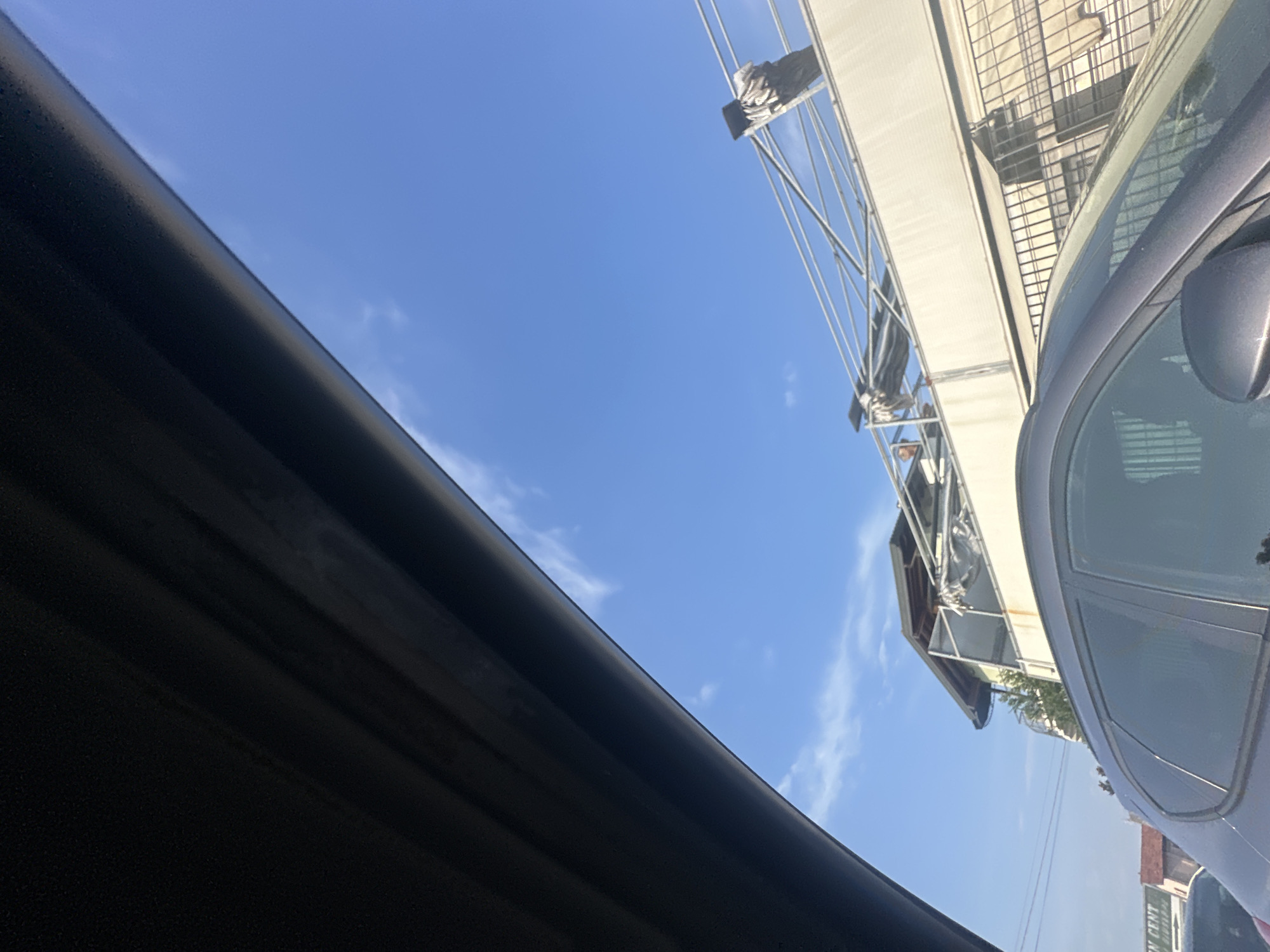 This image, taken from inside a vehicle, shows a view towards a building with a rooftop structure against a bright blue sky with scattered clouds. The foreground is dominated by the interior of the car, with a dark-colored dashboard or headliner extending across the top left portion of the frame. The middle ground features the side of a modern building, characterized by white walls and a tiered roof. Above the building, a metal framework is visible, with some tarps or fabric draped over it. The lower portion of the image shows the rear of a dark-colored car. In the distance, to the left, a sign with the text "IN CENT" is partially visible, suggesting a commercial area. The scene appears to be an urban environment, potentially in Chiari, Italy, given the context.
