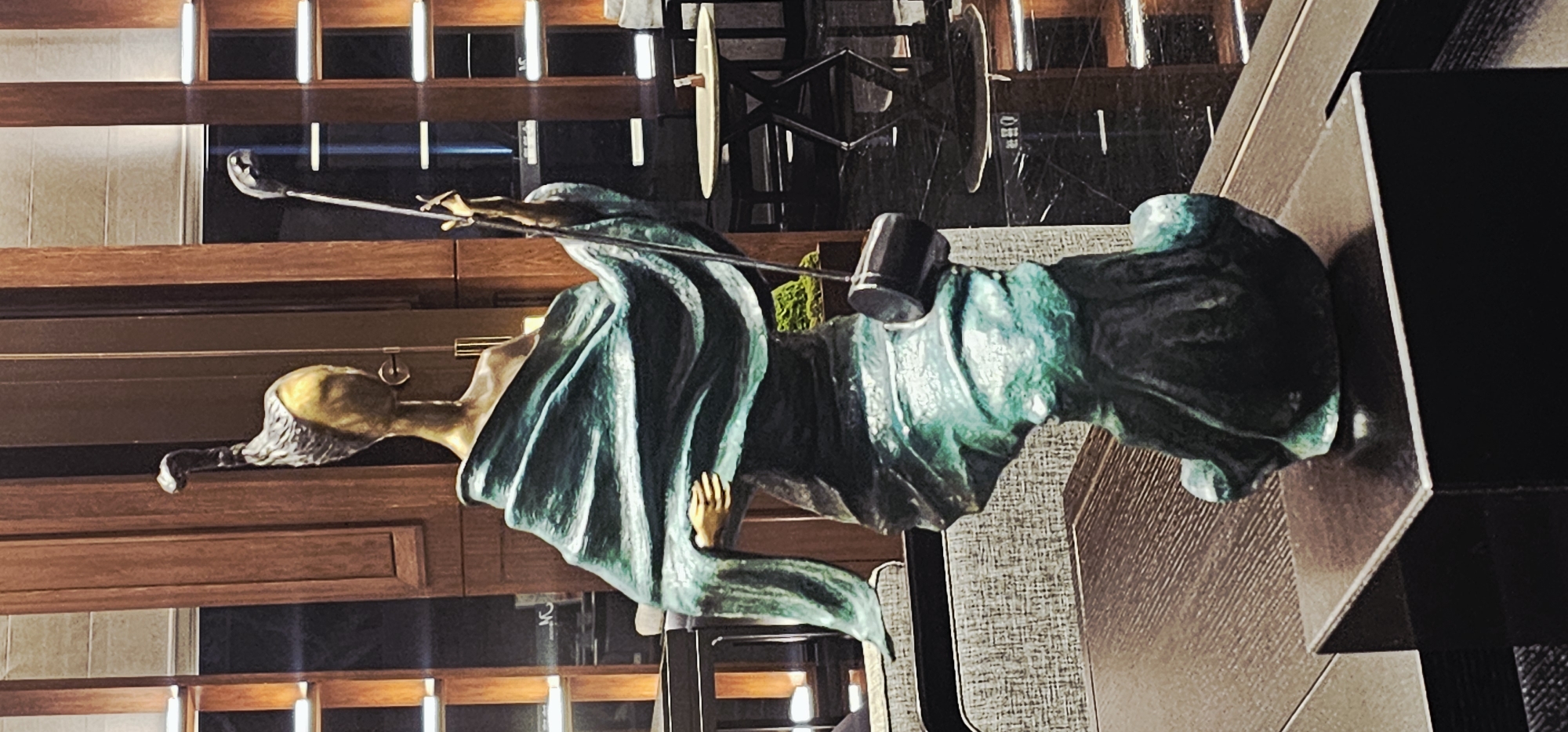 A statue of Lady Justice is displayed inside what appears to be a courtroom or legal office. The statue is made of a bronze-like material with a green patina. The figure wears a draped robe and holds scales in one hand and a gavel in the other. The statue is placed on a table or stand covered with a textured, grey material. The background includes wood paneling and vertical lighting elements. Based on the location given, this image is likely taken in the Liangjiang New Area in Chongqing, China, though no specific landmarks or local elements that would confirm this are visible.