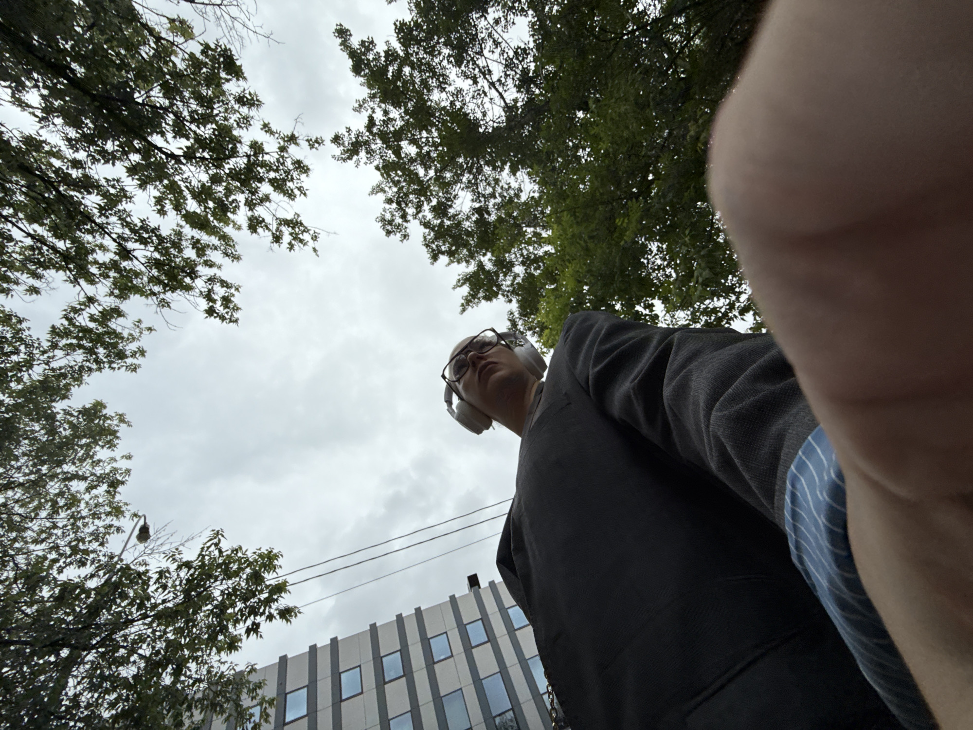 The media file captures a low-angle, wide shot of a person wearing headphones and glasses, looking upwards. The subject is dressed in a dark-colored jacket and appears to be walking outdoors. Overhead, leafy green trees frame the sky, which is overcast with a cloudy, gray appearance, suggesting a daytime setting with diffused light. In the background, a modern building with a facade of white vertical slats and rectangular windows is visible, along with utility wires and a street lamp. The perspective of the shot, with the camera held close to the subject's face and pointing upwards, gives a sense of the environment as perceived by the person. The overall mood is casual and observational, with the focus on the subject's interaction with their surroundings. There is no visible text in the image.