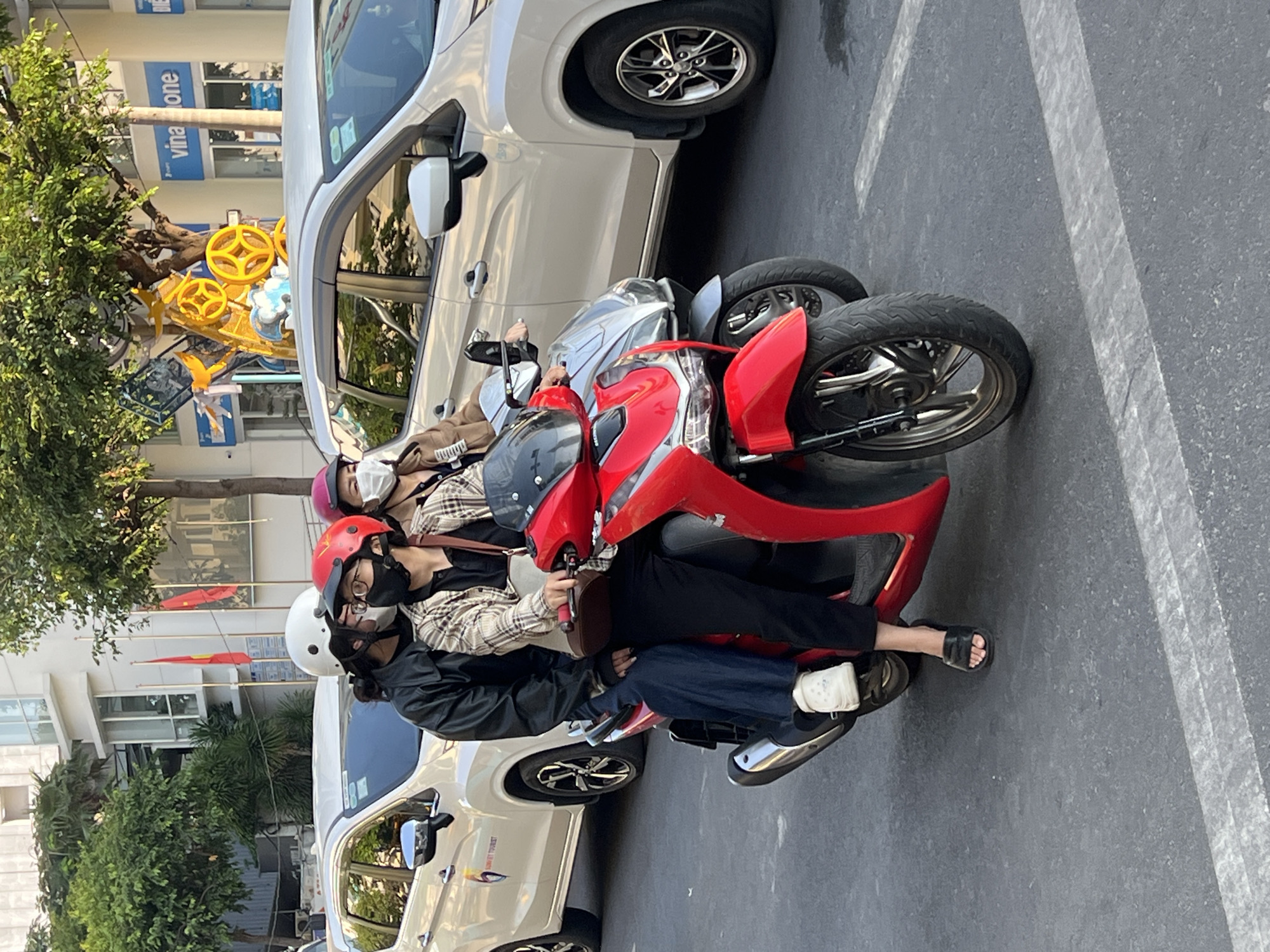 In Nha Trang, Vietnam, a red and silver scooter is parked on a grey asphalt street next to a white car. Three people are on the scooter, all wearing helmets and face masks. The driver is wearing a red helmet and glasses. The person behind them is wearing a pink helmet and a tan jacket. The person in the back is wearing a white helmet and a black jacket. The street has white lane markings. Trees and buildings are visible in the background, including a building with "vina one" signage. Vietnamese flags are flying in front of the building. Decorations with yellow and blue elements are attached to a tree.