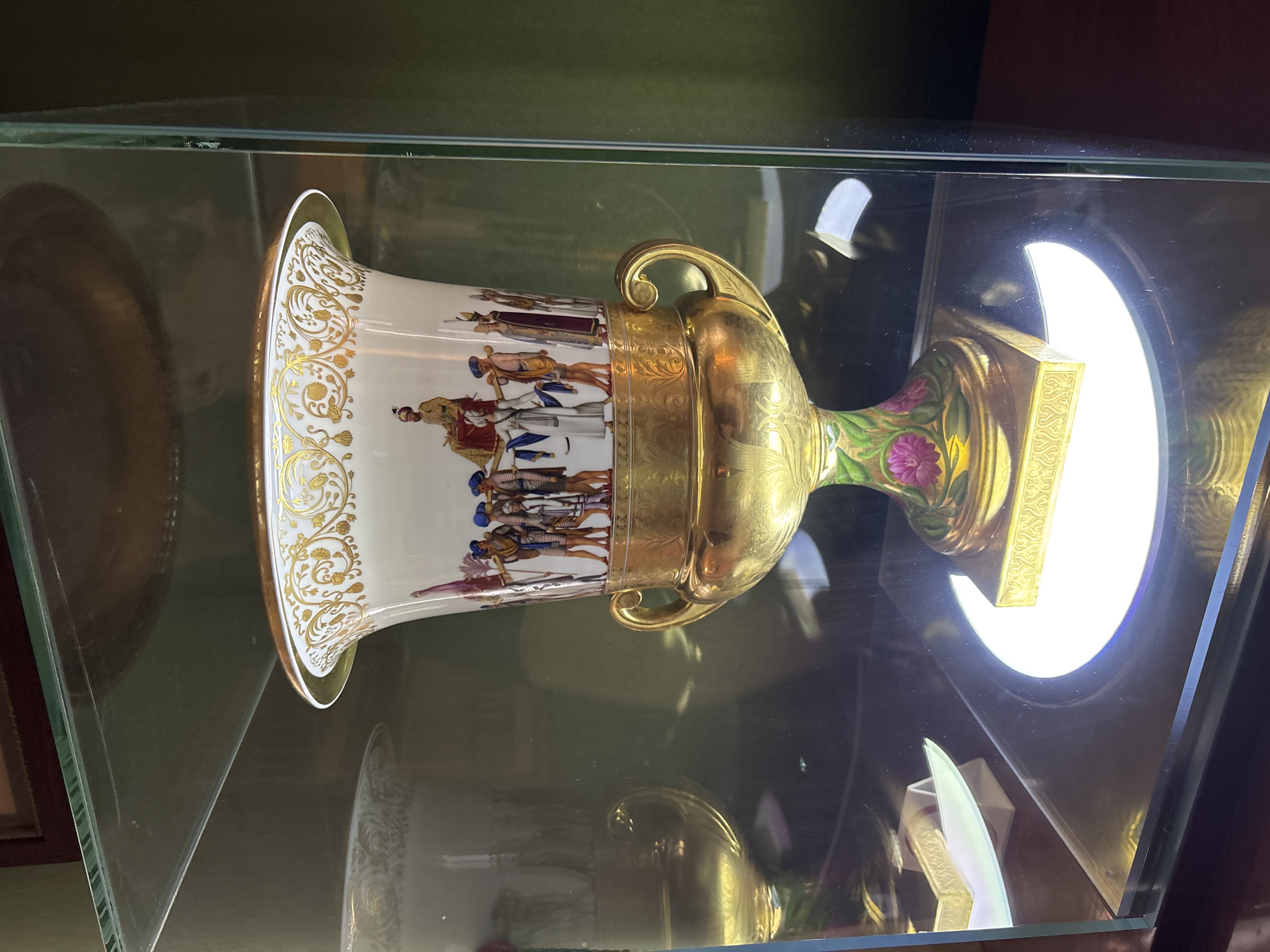 The image shows a detailed, ornate porcelain vase with gold accents and painted figures displayed within a glass case. The vase has a wide, flared opening with a gold rim, decorated with intricate gold scrollwork. Below this, a band of painted figures depicts a procession, with individuals in colorful attire and head coverings, some carrying banners or spears. The central figure appears to be seated on a palanquin. The body of the vase transitions to a gold stem with two ornate handles, leading to a bulbous lower section adorned with painted floral motifs in pink and green. The vase rests on a gilded base, which is illuminated from below by a circular light.
This vase is located in the Great Gatchina Palace in Gatchina, Russia, specifically within the ceremonial rooms or museum exhibition areas. The background consists of a dark green wall, and reflections on the glass case reveal hints of other objects and possibly interior architectural details of the palace. The lighting suggests an indoor display setting, emphasizing the object's artistic and historical value.