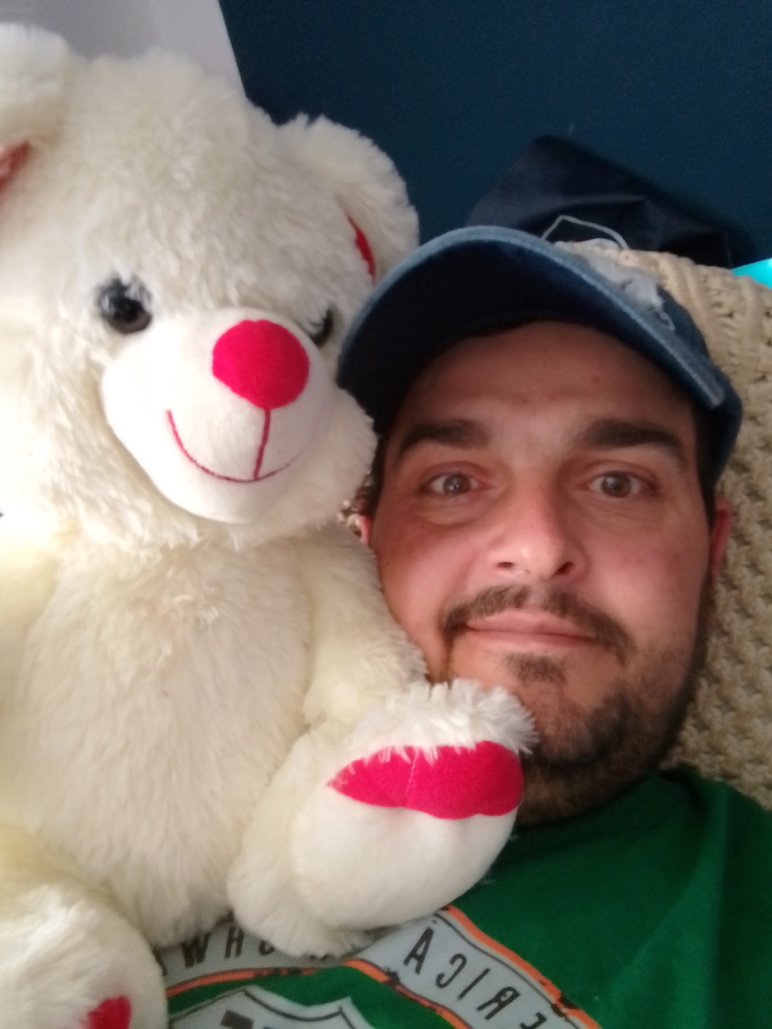 A man is taking a selfie with a white teddy bear. The man is wearing a green t-shirt and a blue baseball cap. He has a beard and mustache. The teddy bear is large and fluffy with a red nose and red accents on its paws. They are both positioned in the foreground of the image. The background is out of focus and appears to be a textured surface and a dark blue wall. There is no information in the image to indicate a specific location in Wedel, Germany.