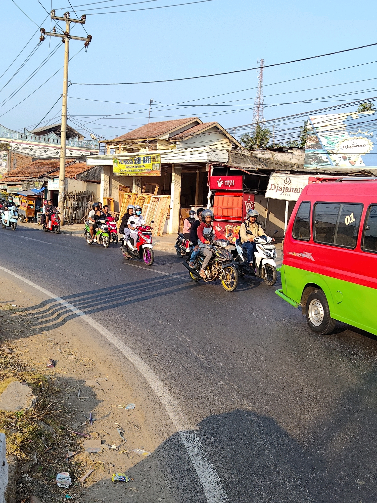 The image depicts a street scene in Purwakarta, Indonesia, likely during the daytime. A procession of motorcycles, each carrying one or two riders, travels along a paved road. Most riders are wearing helmets, and some are also wearing masks. The motorcycles vary in color and style, with some appearing to be automatic scooters and others more traditional models. In the background, there are buildings with tiled roofs, shops, and a prominent utility pole with numerous wires. A red and green van is also visible, partially obscuring some of the motorcycles. The road surface shows signs of wear, and there are shadows cast across it, indicating the sun is at a moderate angle. The environment appears somewhat cluttered, with some debris visible on the side of the road. A tall communications tower can be seen in the distance.