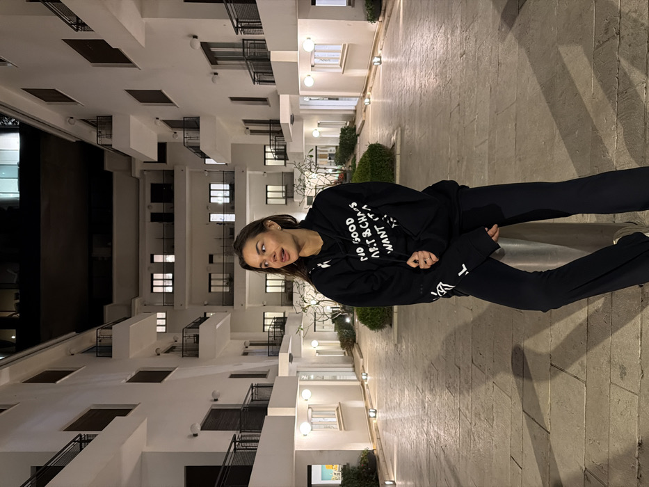 A young adult female is positioned outdoors at night in a paved courtyard within a residential or commercial complex. She is leaning against a cylindrical silver bollard, facing slightly left while looking towards the right of the frame. She wears a black hooded sweatshirt with white text partially visible on the front ("NO GOOD ART & CHAOS I WANT IT ALL"), black flared trousers, and light-colored athletic shoes. Her hands are resting in front of her. The background consists of multi-story, white-facaded buildings with multiple windows, some illuminated, and dark balconies with railings. The architectural style exhibits clean lines and functional design, characteristic of Bauhaus or International Style architecture prevalent in Tel-Aviv, Israel. The paved ground is illuminated by recessed ground lights, and wall-mounted sconces provide additional light on the building facades. Manicured shrubbery and small trees are visible in garden beds along the edges of the paved area. The overhead sky is dark.