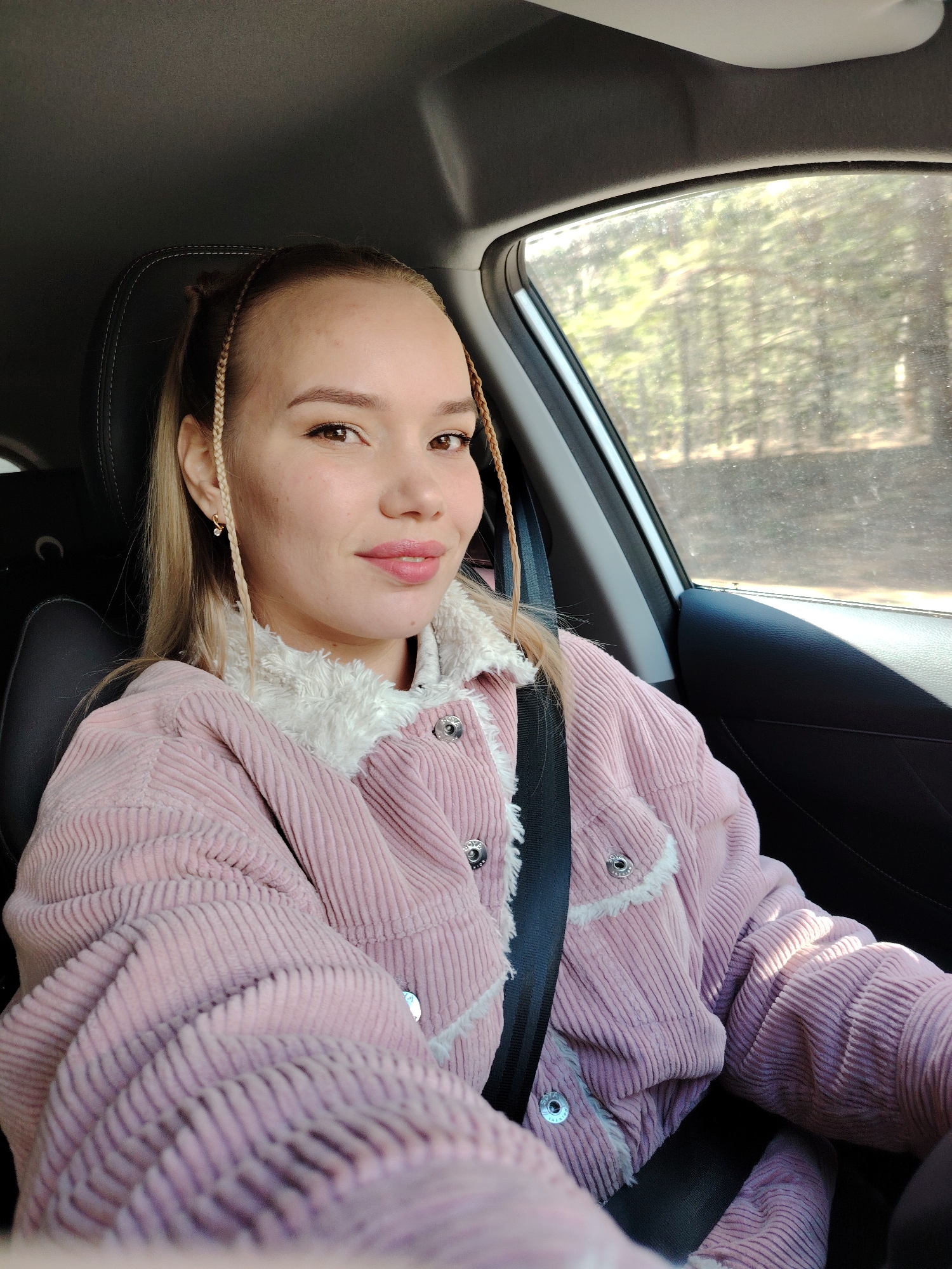 A young woman is seated in the driver's seat of a car, taking a selfie or being photographed. She has blonde hair with two thin braids framing her face, and a soft, pleasant smile on her lips. She is wearing light makeup, including pink lipstick, and small gold-colored earrings. Her attire consists of a textured pink corduroy jacket with a white fluffy collar and trim, secured with silver buttons. A black seatbelt is visible across her chest and lap.
The interior of the car is visible, including a black headrest behind her, a dark car door panel, and part of the steering wheel on the right side of the frame. Through the car window, a blurred background of trees and foliage suggests the vehicle is in a natural setting, possibly moving along a road in or near Chaikovsky, Russia. The bright light from outside indicates it is daytime and sunny. There is no visible text in the image.