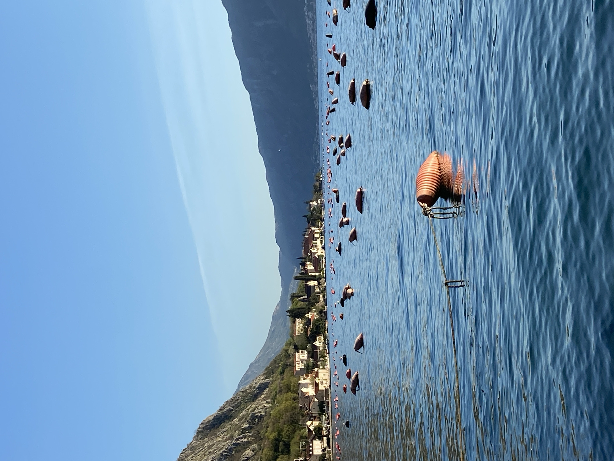 The image depicts the town of Donji Orahovac, Montenegro, situated on the Bay of Kotor, with a mountain range in the background. The foreground shows a body of water, possibly the bay, with numerous orange buoys floating in it. Some of the buoys appear to be connected by ropes or lines. The sky is clear and blue. The town itself consists of buildings with light-colored roofs and is nestled between the mountain and the shoreline.