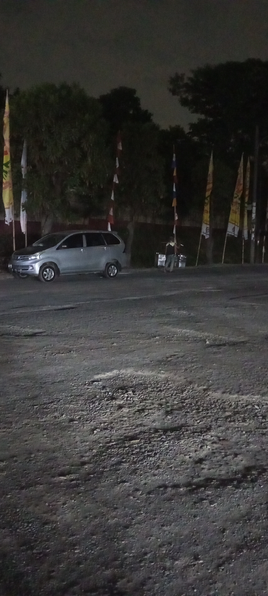 The image depicts an outdoor scene at night, likely in a parking area or along a road. A silver minivan is parked on the left side of the frame, facing towards the right. The vehicle appears to be a modern passenger car. In the background, there are several trees casting shadows and a line of flags on poles, some of which are white with red accents, and others are colorful with various designs. A person is visible on the right side of the frame, near a small structure or cart, wearing dark clothing. The ground is paved with an uneven surface, appearing to be asphalt or gravel with visible patches and debris. The sky above is dark, indicating nighttime, with some faint atmospheric glow. The overall impression is a low-light, possibly urban or semi-urban environment.