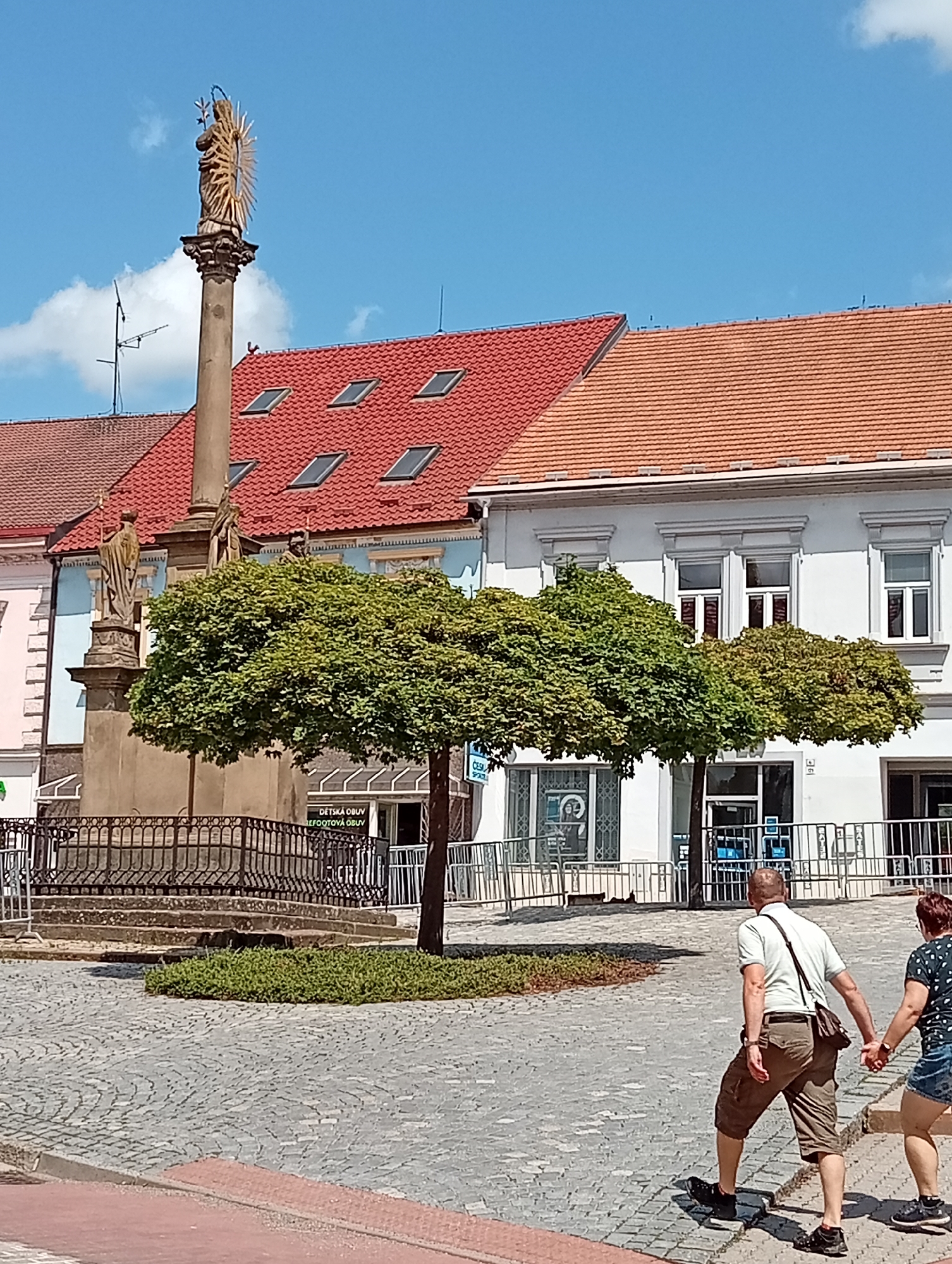 In the city of Tišnov, Czechia, a sunny day unfolds in a town square paved with cobblestones. A stone monument, topped with a golden-haloed statue, stands prominently, flanked by two smaller stone figures at its base. In the foreground, a pair of figures, a man and a woman, walk hand-in-hand across the cobblestones. The man, wearing a light-colored polo shirt and brown shorts, carries a dark bag slung across his shoulder. The woman, dressed in a patterned top and denim shorts, walks beside him. Behind the monument, a small, well-maintained green bush encircles the base of a tree with a rounded, dense canopy. Beyond the monument and trees, a row of buildings lines the square. The building on the left has a red tiled roof with multiple dormer windows, while the building to its right features an orange tiled roof and a white facade with decorative window frames. Shops with glass storefronts are visible, and temporary metal barriers are erected in front of some of them. The sky above is a clear, bright blue with a few scattered white clouds. The overall scene depicts a pleasant day in a European town square, with tourists or residents enjoying the public space.