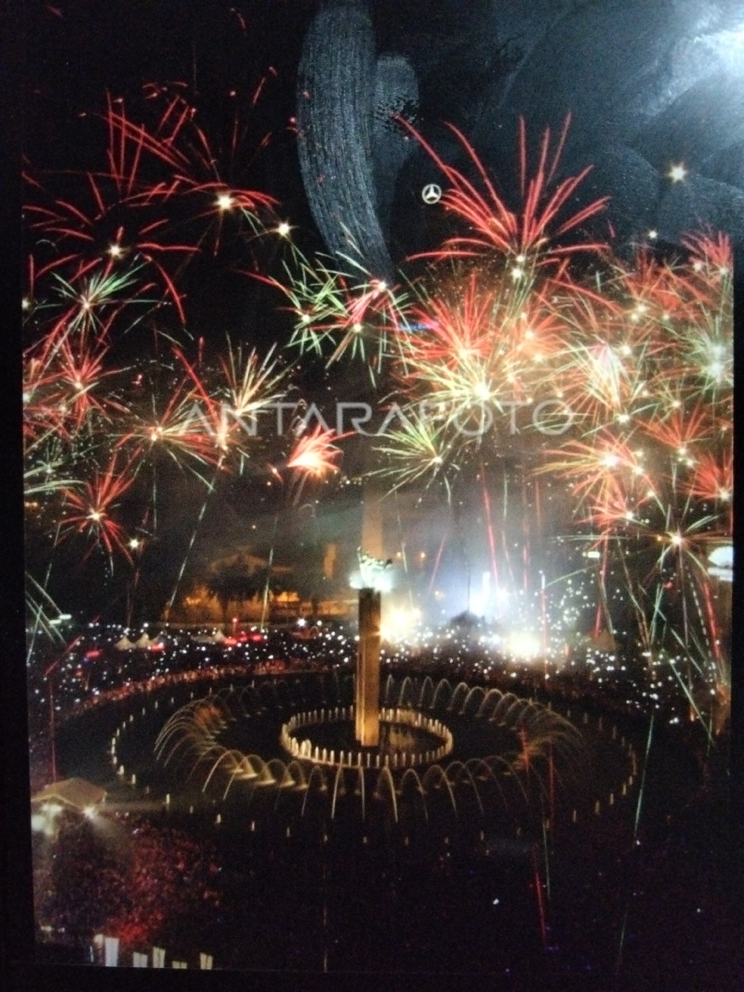 The image depicts a nighttime fireworks display over a large crowd gathered around a fountain in what appears to be a city park. The fireworks are bursting in vibrant colors of red, green, and yellow, illuminating the dark sky. A large, illuminated monument stands tall in the center of the fountain area, providing a focal point. The crowd below is a sea of people, many holding up lights or phones, indicating they are actively watching the event. The fountain itself is a prominent feature, with arcs of water illuminated by lights. The overall scene suggests a celebratory public event, possibly a festival or national holiday, taking place in Kualaraja, Indonesia, given the presence of the ANTARAPOTO watermark, indicating an Indonesian news agency.
