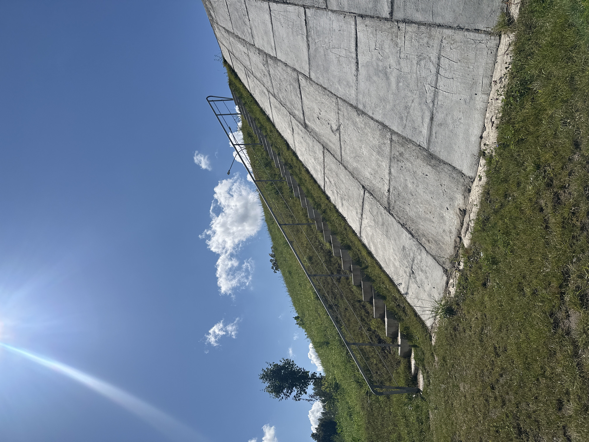 An outdoor, low-angle, eye-level shot captures a concrete embankment and a grassy hill under a bright blue sky with scattered clouds and a strong sun glare in the upper left corner. A metal handrail runs diagonally up the side of the embankment, alongside a set of concrete stairs. The embankment is partially covered with grass and the lower section is a smooth concrete surface, divided into large rectangular panels. A single, medium-sized deciduous tree stands on the grassy hill. The scene appears to be a public space, possibly a park or a waterfront area, in Minsk, Belarus. There are no people or animals visible in the image.