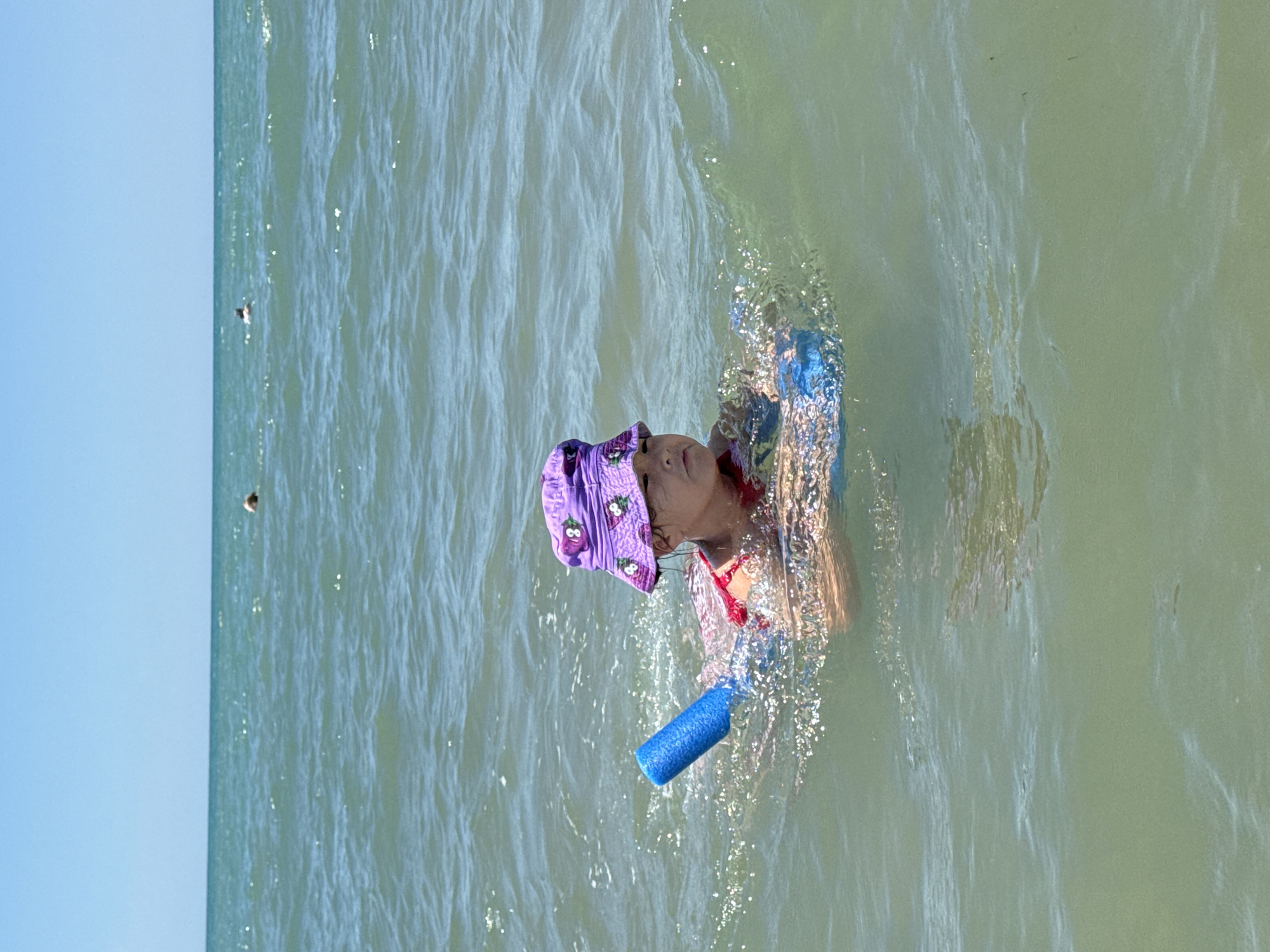 A young child, wearing a purple bucket hat with a floral pattern and a red swimsuit, floats in the sea, holding onto a blue foam pool noodle and a kickboard. The child is looking to the right with a neutral expression. The water is a greenish-blue color, with small waves and sparkling reflections of the sun. In the background, two distant figures are visible swimming in the ocean under a clear blue sky. The location is the city of Prostorny, Russia.