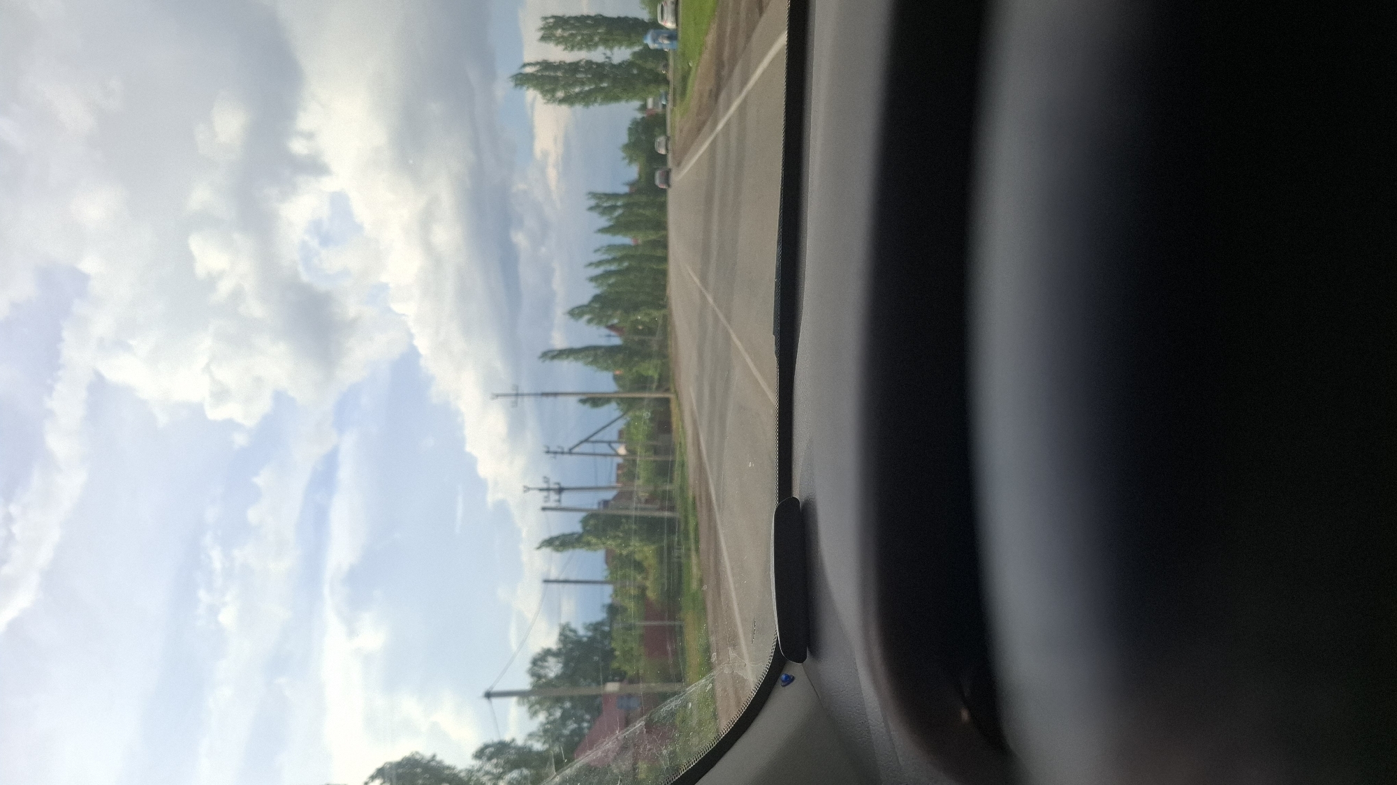 The image depicts a daytime view from inside a vehicle looking forward through the windshield onto a rural or semi-rural road in Прибрежный, Russia. The sky occupies the upper portion of the frame, showing a mix of large white and grey cumulus clouds against patches of light blue sky.
The central feature of the image is an asphalt road, marked with a continuous white line down its center, suggesting a two-lane configuration (one lane per direction). The road appears to extend straight into the distance, converging at the horizon. In the far distance on the road, several indistinct passenger vehicles are visible.
Along the left side of the road, a series of wooden and metal utility poles with overhead power lines are visible. Beyond these poles, obscured by green foliage, are residential-style buildings with reddish roofs. A fence runs along the roadside, separating the properties from the road. On both sides of the road, a distinct line of tall, slender, columnar trees (possibly poplars) extends into the distance. On the far right side of the road in the distance, two light-colored structures, resembling portable toilets, are present.
The lower half of the image is dominated by the interior of the vehicle. A dark, textured dashboard is prominent, with an elongated dark object mounted near the base of the windshield, possibly a dashcam or navigation device. A small blue clip or accent is visible on the dashboard to the left. The left A-pillar and a portion of the vehicle's interior trim are also visible. The windshield shows some minor dust or streaks. No people are explicitly visible in the scene, either inside or outside the vehicle.