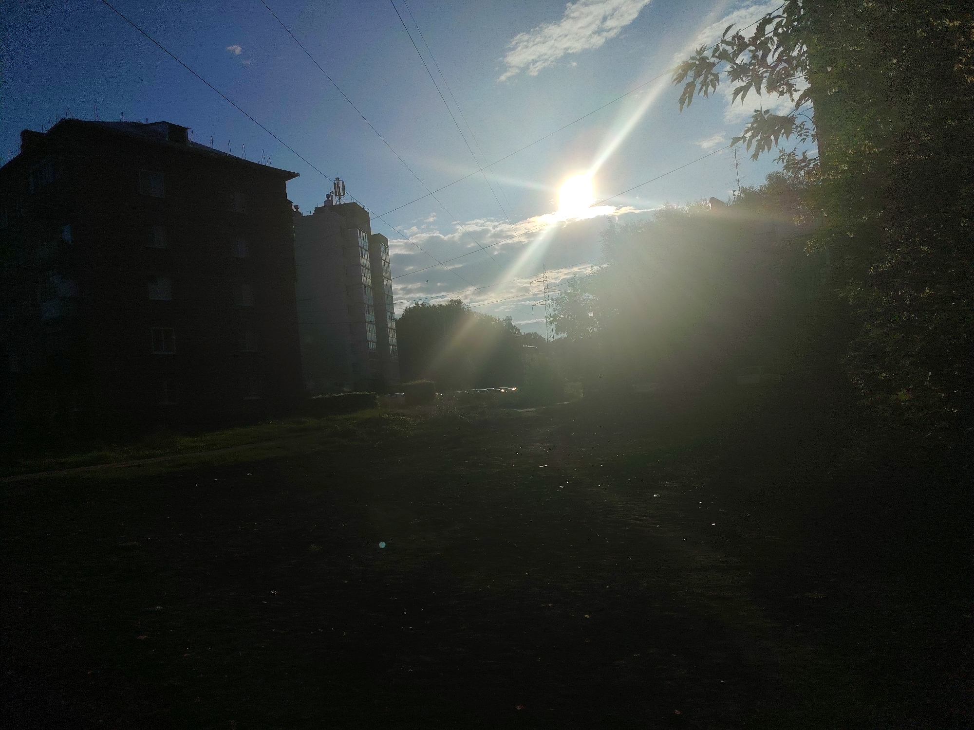 The image captures a view of two apartment buildings in the city of Kemerovo, Russia, under a bright, midday sun. The sun, positioned high in the sky, is the primary light source, creating a strong glare and lens flare that washes out parts of the image. The sky is a clear blue, with scattered white clouds. Overhead, power lines crisscross the frame, indicative of urban infrastructure.
The apartment buildings are silhouetted against the bright sky, making their details difficult to discern. The building on the left appears to be a darker, possibly brick structure, with multiple floors and windows. The building on the right is lighter in color and seems to have a communication antenna or tower on its roof.
In the foreground, a grassy area is visible, much of it cast in deep shadow due to the sun's position. The dark ground suggests it might be a courtyard or a park area adjacent to the buildings. Trees with lush green foliage are present on the right side of the image, partially obscuring the view and adding to the overall contrast. The overall impression is a scene of everyday urban life in Kemerovo, characterized by the presence of residential buildings, power infrastructure, and natural elements, all illuminated by the strong, direct sunlight.