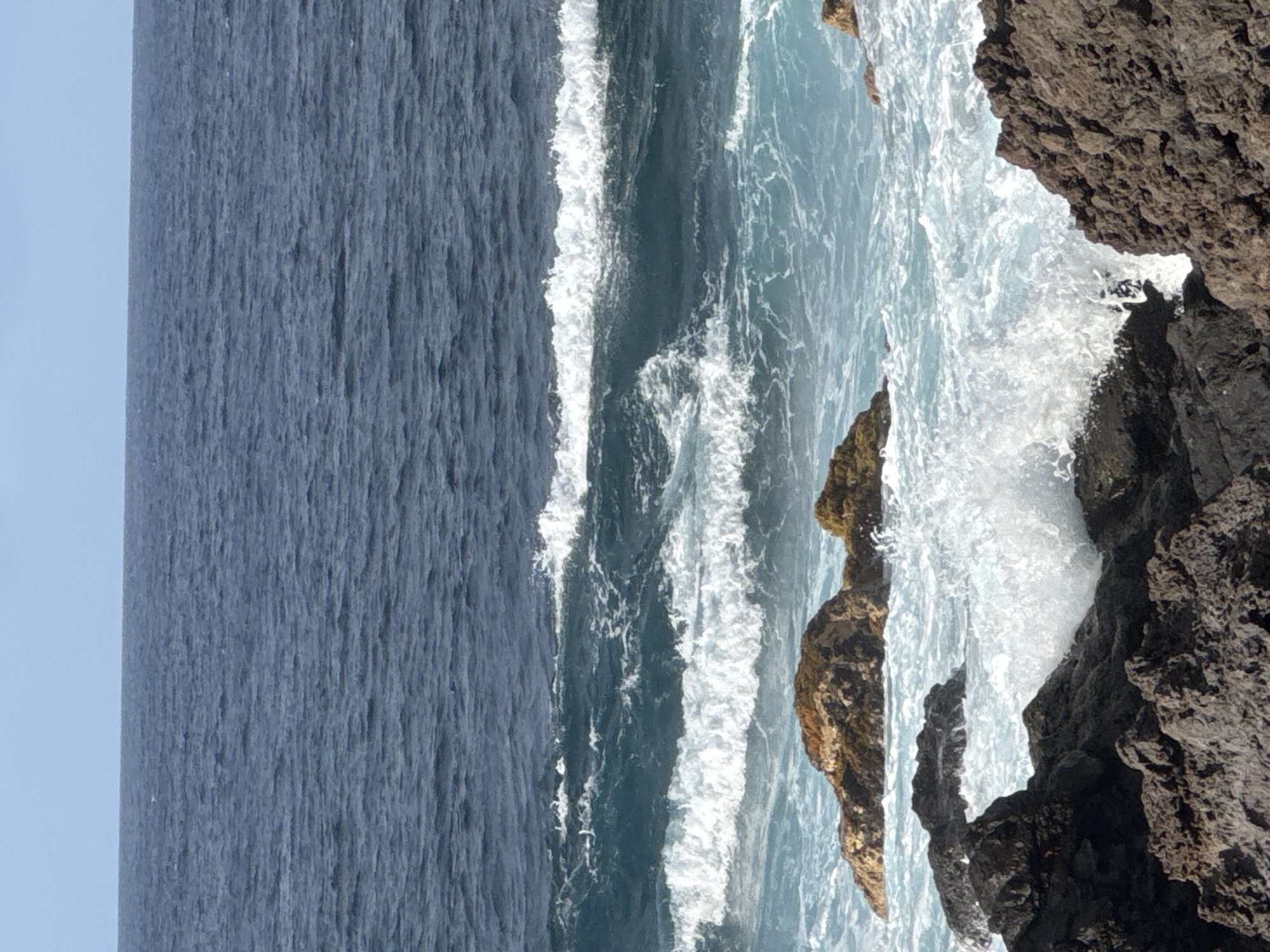 The image shows a view of the ocean from a rocky coastline. The sea is a deep blue and is moderately wavy, with white foam cresting the waves. There are several dark, rugged rocks in the foreground, with some lighter-colored, moss-covered rocks further out in the water. The sky is a pale blue. There are no people or man-made structures visible in the image.