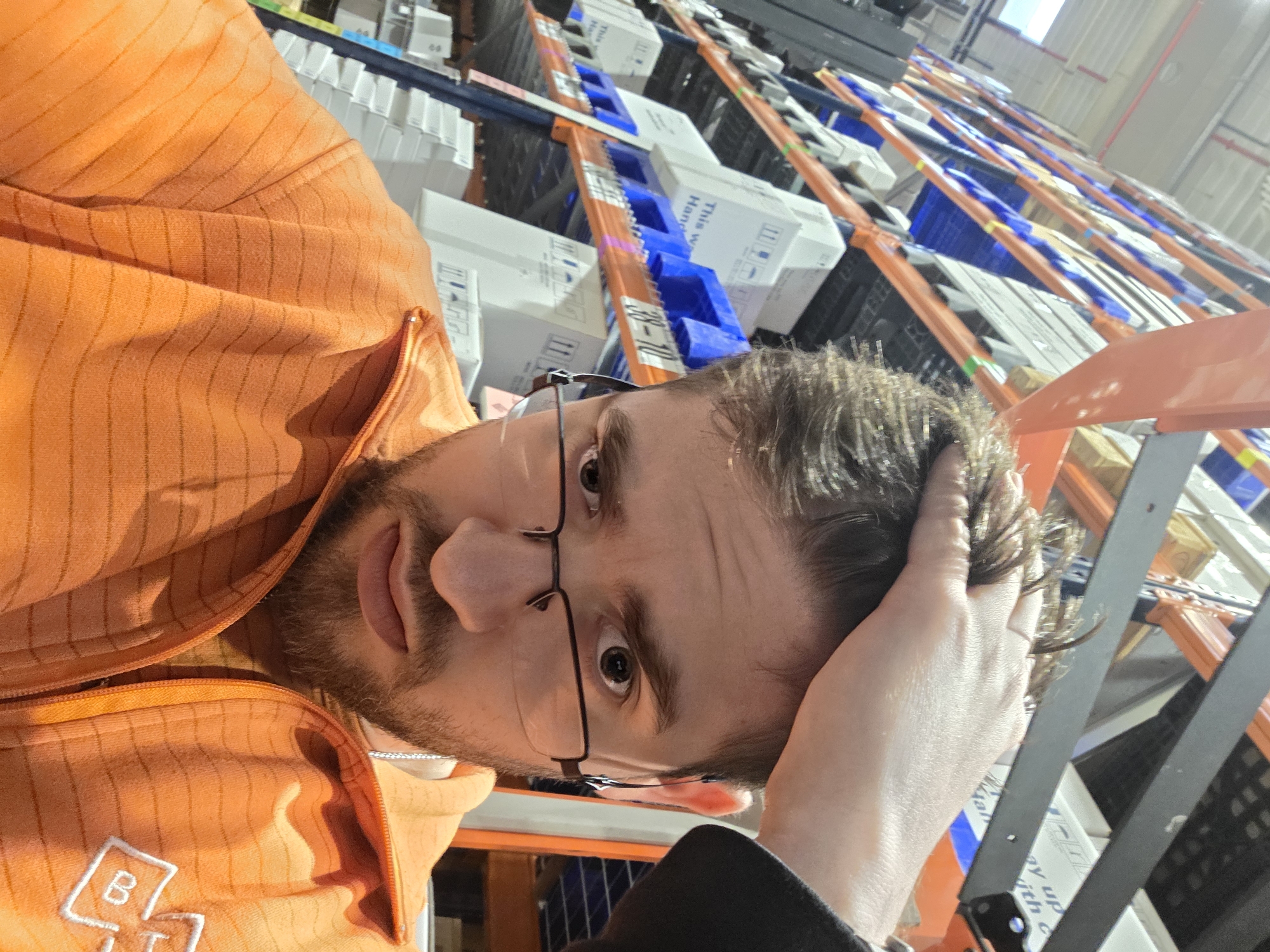 A man wearing glasses and an orange sweatshirt is in a warehouse. He has his hand on his head. Behind him are rows of shelving units filled with boxes and blue plastic containers. The location is in Plovdiv, Bulgaria.