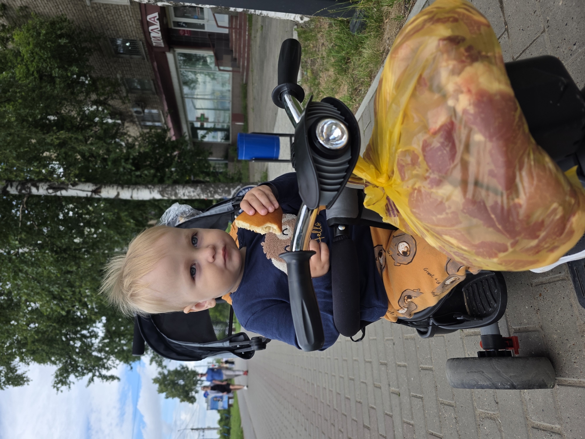 A young Caucasian boy with blond hair is seated in a black stroller. He is wearing a navy blue long-sleeved shirt and orange pants with a pattern of bears. The boy is holding a piece of bread and appears to be eating it. The stroller has black handlebars with a headlight and a yellow bag filled with raw meat is attached to the front. The stroller is on a paved walkway in what appears to be a city. In the background, there are trees, a brick building with windows and a sign that reads "МИЛА" (MILA), and a blue trash can. There are also two adults walking in the distance on the walkway. The overall scene is outdoors on a cloudy day in Chaikovsky, Russia.
