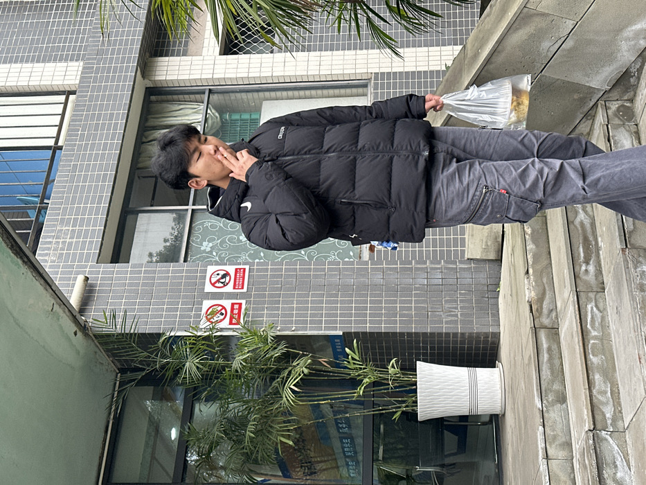 A young man stands outside a building. He's wearing a black puffer jacket and grey pants, holding a clear plastic bag in one hand and appears to be smoking. The building has windows, a no-feeding-animals sign, and potted bamboo in front. The building's exterior is a mix of grey brick and concrete. The area where the man is standing is paved with grey stone. The picture is taken at an angle.