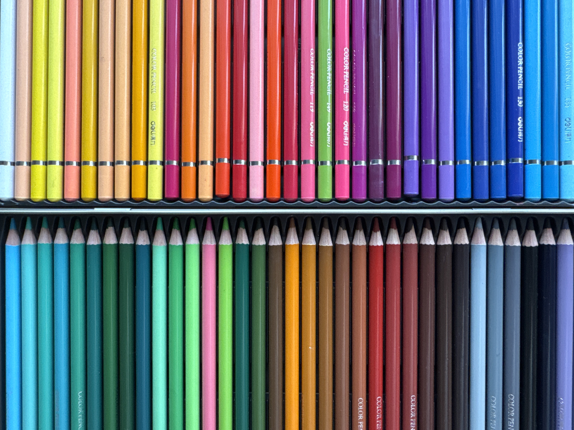 The image is a top-down, close-up shot of a tin containing rows of colored pencils. The pencils are arranged horizontally in two columns within the tin, with the tips pointing towards the right side of the frame. The colors are vibrant and organized from light blue and green on the left column, transitioning through shades of green, brown, orange, and red. The right column starts with yellow and pink, then moves through orange, red, pink, green, purple, and blue. Some of the pencils have text printed on them, indicating they are "COLOR PENCIL" and some numbers like "103", "117", "149", "120", "130", and "136". The background is the interior of the tin, which appears to be black. There are no people, animals, or any elements related to the city of Nha Trang Ward, Vietnam present in the image.