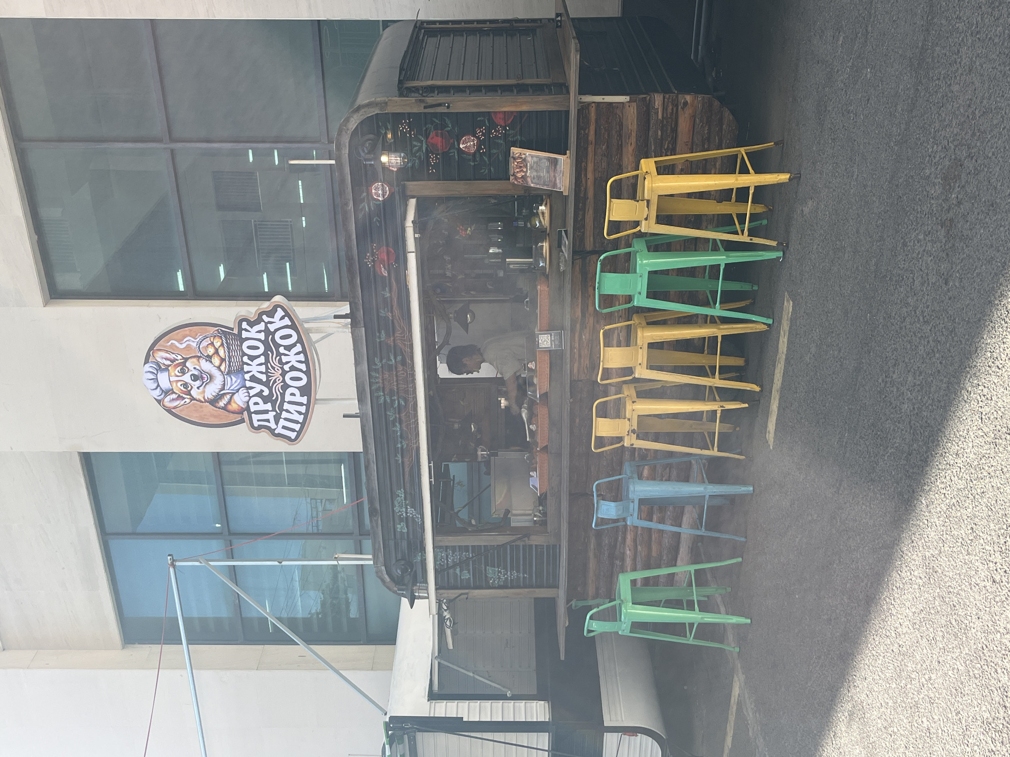 The image shows a food stand in Bac Nha Trang Ward, Vietnam, with a sign that reads "ДРУЖОК ПИРОЖОК" featuring a dog wearing a chef's hat. The food stand is constructed from dark metal and wood, with a floral design on its upper portion. Inside the stand, a person wearing a white shirt is visible. Several stools in various colors – yellow, green, and light blue – are arranged along the side of the stand. A large building with a glass window façade is behind the stand, and an awning extends partially from the building's front. The ground is paved with asphalt.