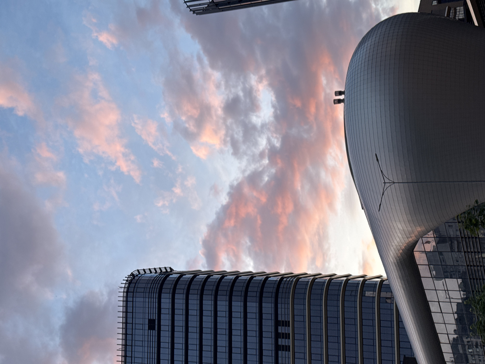 The image presents a modern urban landscape in Minsk, Belarus, at either sunrise or sunset. The sky, occupying the upper two-thirds of the frame, displays scattered cumulus clouds illuminated with pink and orange hues against a pale blue background, indicative of low sun angles. On the left, a prominent multi-story building features a dark, reflective glass facade with rounded corners and horizontal banding. A series of vertical fin-like structures are visible along its curved roofline. This structure is identified as one of the Dana Towers within the Mayak Minska complex. In the lower-center and right, a distinct, large architectural structure with a smooth, continuously curved, panelized metallic exterior, resembling an elongated dome or shell, dominates the foreground. Two dark, cylindrical elements, likely vents or decorative features, are positioned atop this curved building. This structure is identified as the Capital Palace, also within the Mayak Minska complex. A dark-colored street light pole with an extended arm is visible in front of the Capital Palace. Reflections of distant urban elements and sky are evident in the glass surfaces of both buildings. No human figures are discernible.