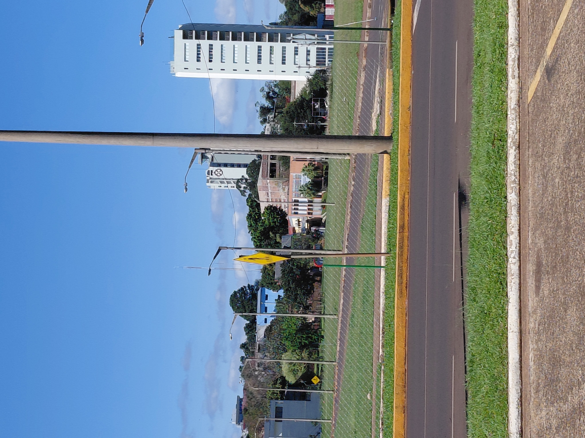 The image shows a street scene in Encarnacion, Paraguay, on a clear day with a bright blue sky and scattered clouds. In the foreground, a paved road with white lane markings is visible, bordered by a strip of green grass and a yellow curb. Beyond the road, a chain-link fence encloses a grassy area with several lampposts.
In the background, a cluster of buildings is present, including a tall, modern white high-rise building with many windows, and another taller building with a circular design near its top. Several smaller buildings, some appearing to be industrial or office spaces, are also visible, along with lush green trees and vegetation. A yellow diamond-shaped sign with a pedestrian silhouette is mounted on a pole near the fence. A large, light-colored utility pole stands prominently in the middle ground.