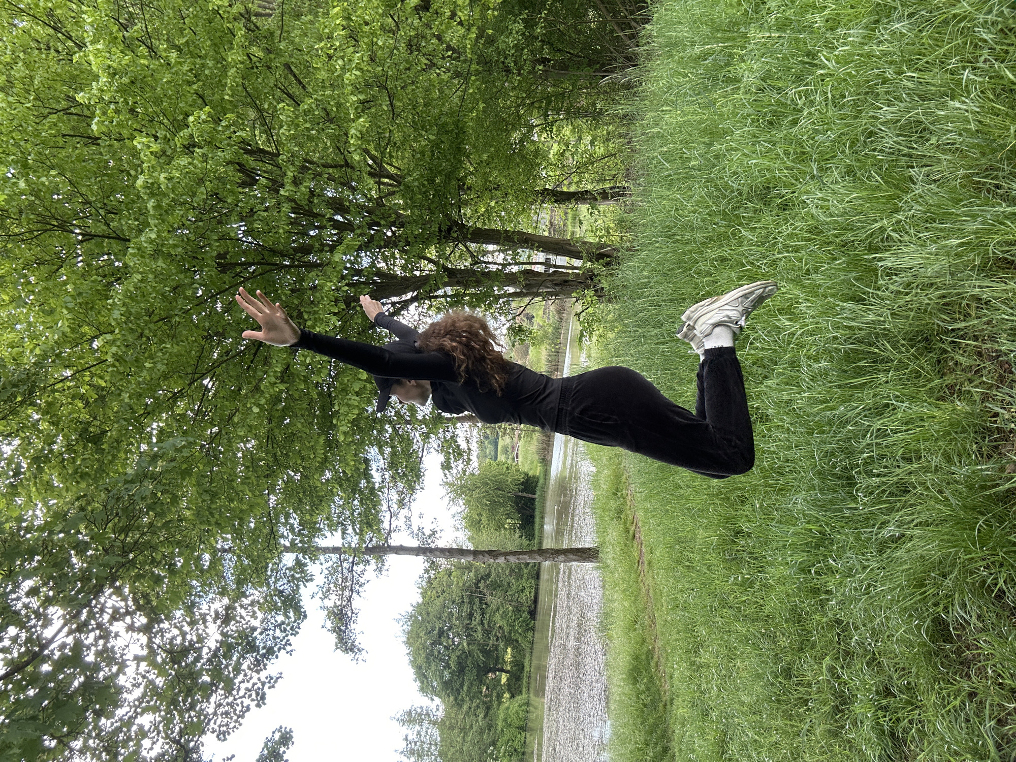 A woman in a black tracksuit and sneakers is standing in a field in Flein, Germany. Her arms are outstretched and she is looking up towards the trees. The field is green and lush, with tall grass. The sky is cloudy. There is a body of water in the background.