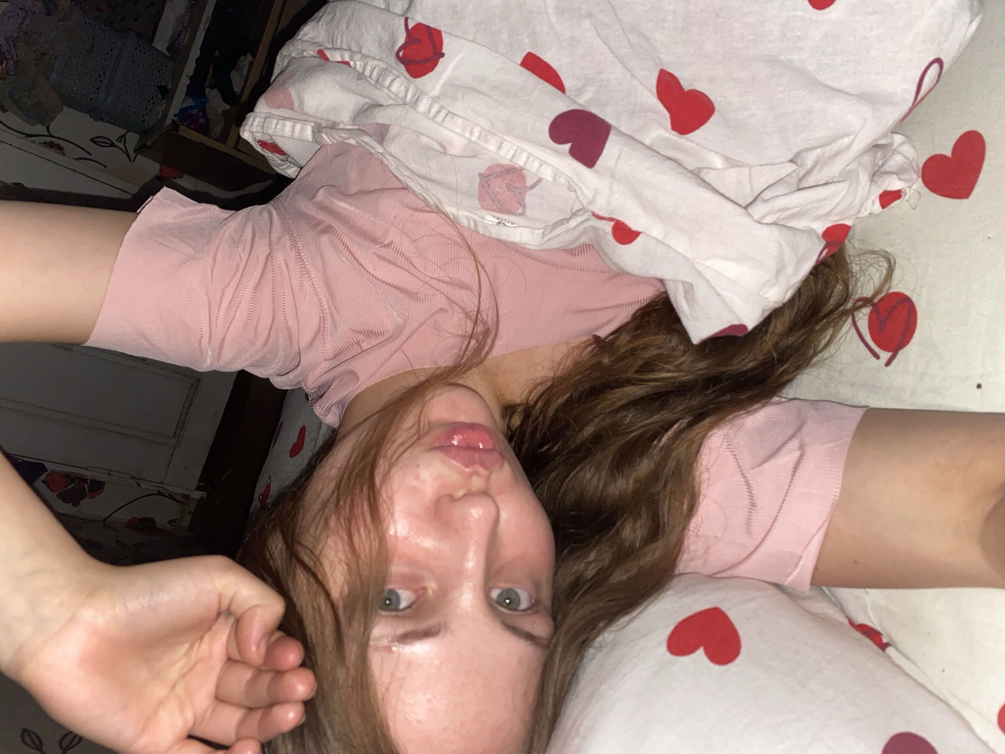 A young woman with long, brown hair is lying down, possibly in bed. She is wearing a pink top and is partially covered by a white blanket with red and pink heart patterns. Her left arm is raised above her head, resting on a pillow that also has a heart pattern. Her face is turned towards the camera, and she has green eyes and glossy lips. The background appears to be an indoor setting with hints of furniture and a wall with floral wallpaper. The lighting suggests it might be evening or a dimly lit room. The location of Вассята, Russia, is not evident from the image itself.