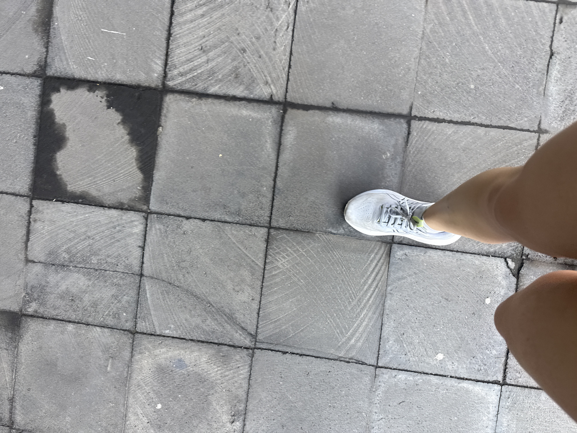 The image depicts a section of a paved sidewalk with grey square tiles. A dark, wet patch is visible on one of the tiles. Part of a person's legs, wearing a white athletic shoe, is present in the frame, suggesting they are standing on the sidewalk. The scene appears to be outdoors, given the lighting and the nature of the paving stones. Given the context of Mexico City, Mexico, the tiles may be part of a public walkway or pedestrian area within the city.