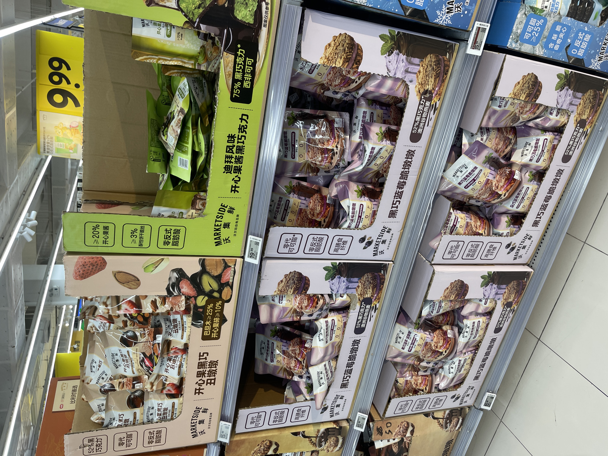 The image shows a retail display of various snack products, primarily cookies and chocolate bars, arranged on store shelves. The products are mostly packaged in colorful boxes and bags, with prominent branding visible. Some of the packaging features images of fruits like strawberries and blueberries, as well as nuts and chocolate. Price tags are visible, indicating that these items are for sale. The overall environment appears to be a supermarket or a large grocery store, with bright overhead lighting and a tiled floor. In the background, other shelves stocked with products are visible, suggesting a typical retail setting. The specific location of Panyu District, China, cannot be determined from the visual information provided in the image itself; it is external context.