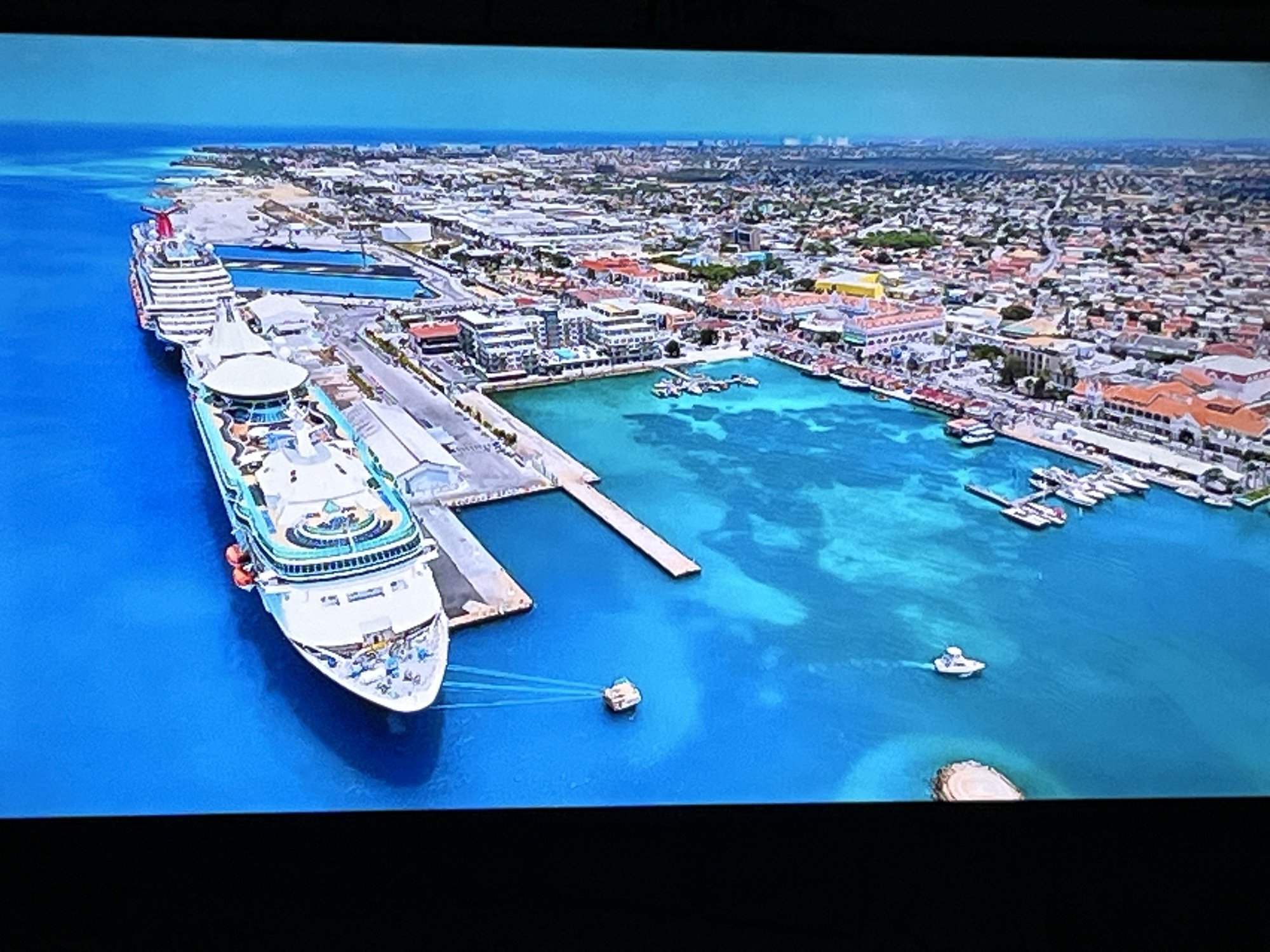 The image is an aerial view of a city with a large cruise ship docked at a port. The city appears to be a coastal town with a densely populated urban area featuring buildings of various sizes and colors. There is a vibrant turquoise body of water in the foreground, with several smaller boats and yachts visible. The cruise ship is white with red and white accents and is docked at a pier. In the background, the ocean extends to the horizon under a clear blue sky. The image does not depict the city of Kharkiv, Ukraine, as Kharkiv is an inland city and not located on the coast.