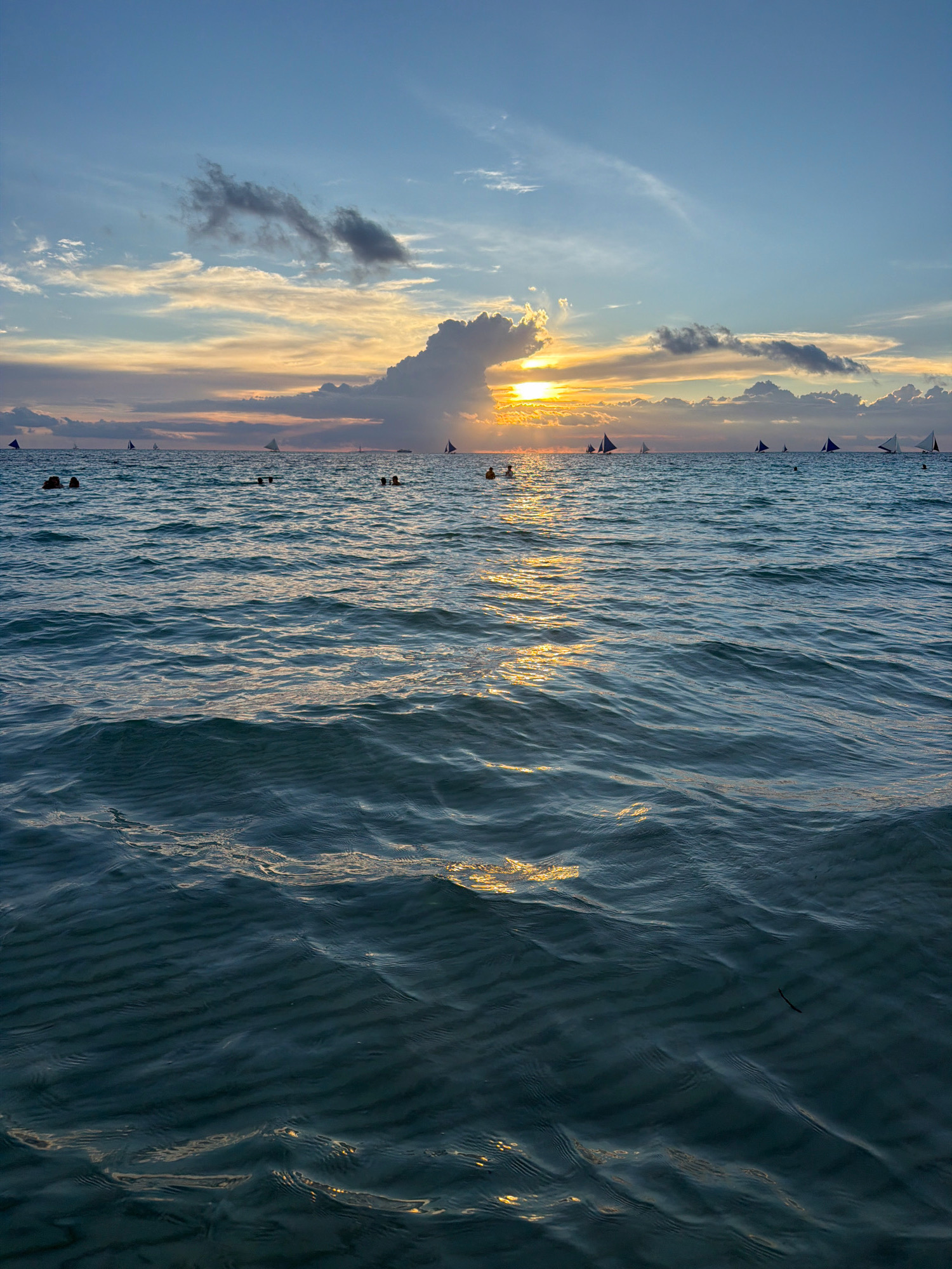 The image captures a serene sunset scene at White Beach in Malay, Philippines. The sky displays a gradient of blues and yellows, with scattered clouds tinged by the setting sun. A prominent, large cloud formation is illuminated by the sun's rays, casting a warm glow. The ocean is a tranquil turquoise, with gentle ripples reflecting the colors of the sky and the sun. In the distance, several sailboats with triangular sails are visible, dotting the horizon. A few people are swimming or wading in the shallow waters closer to the foreground, their figures appearing as silhouettes against the bright ocean surface. The overall ambiance is peaceful and picturesque, characteristic of a tropical beach at dusk.