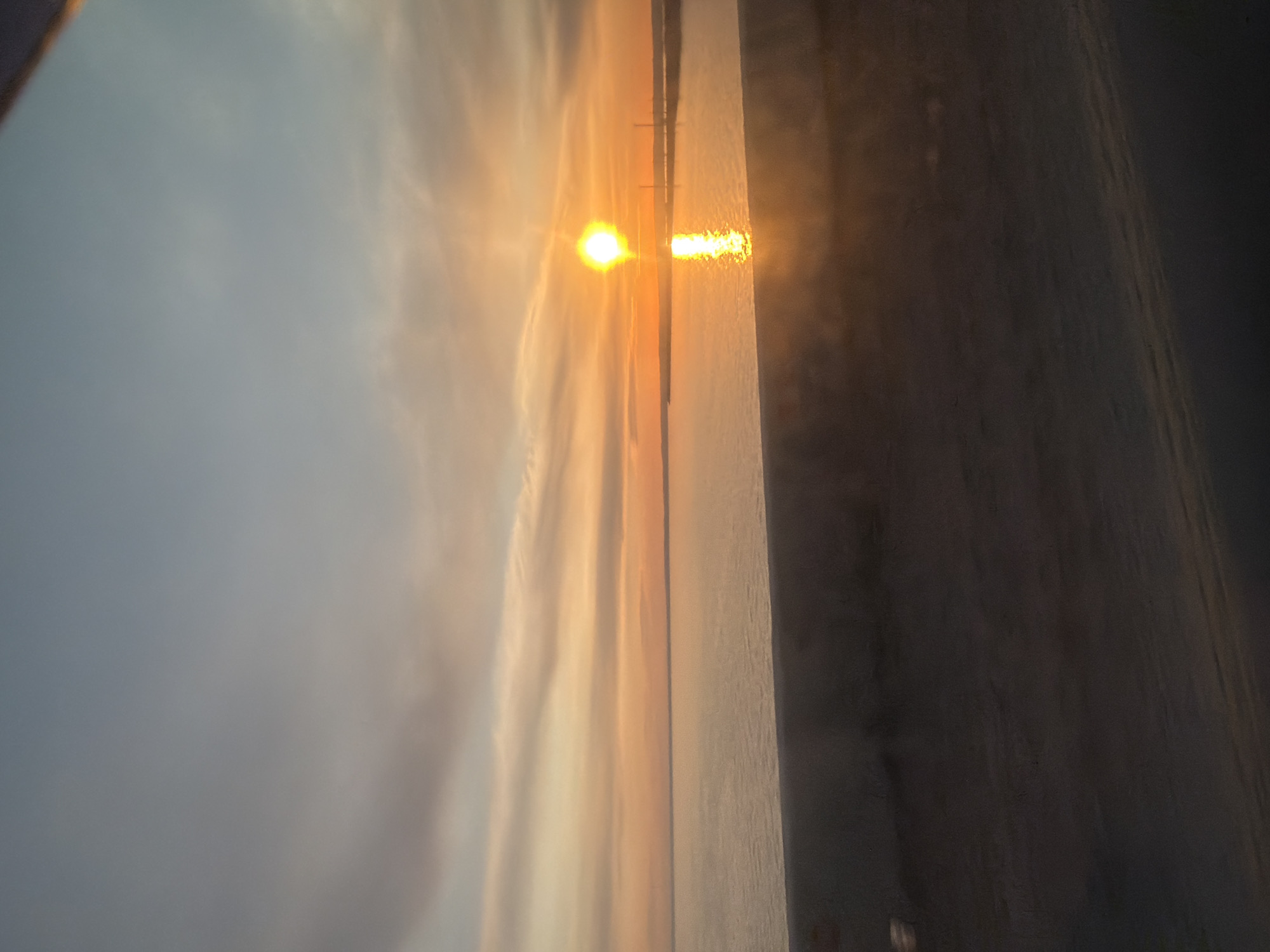 The image shows a sunset reflecting off the water, likely a body of water in Chaikovsky, Russia. The sky is a gradient of colors from pale blue to orange and yellow near the horizon. The sun is a bright orb with its reflection shimmering on the water's surface. A vertical structure appears on the right, possibly a wall or building, that is partly obscured. The overall scene has a hazy, atmospheric quality.