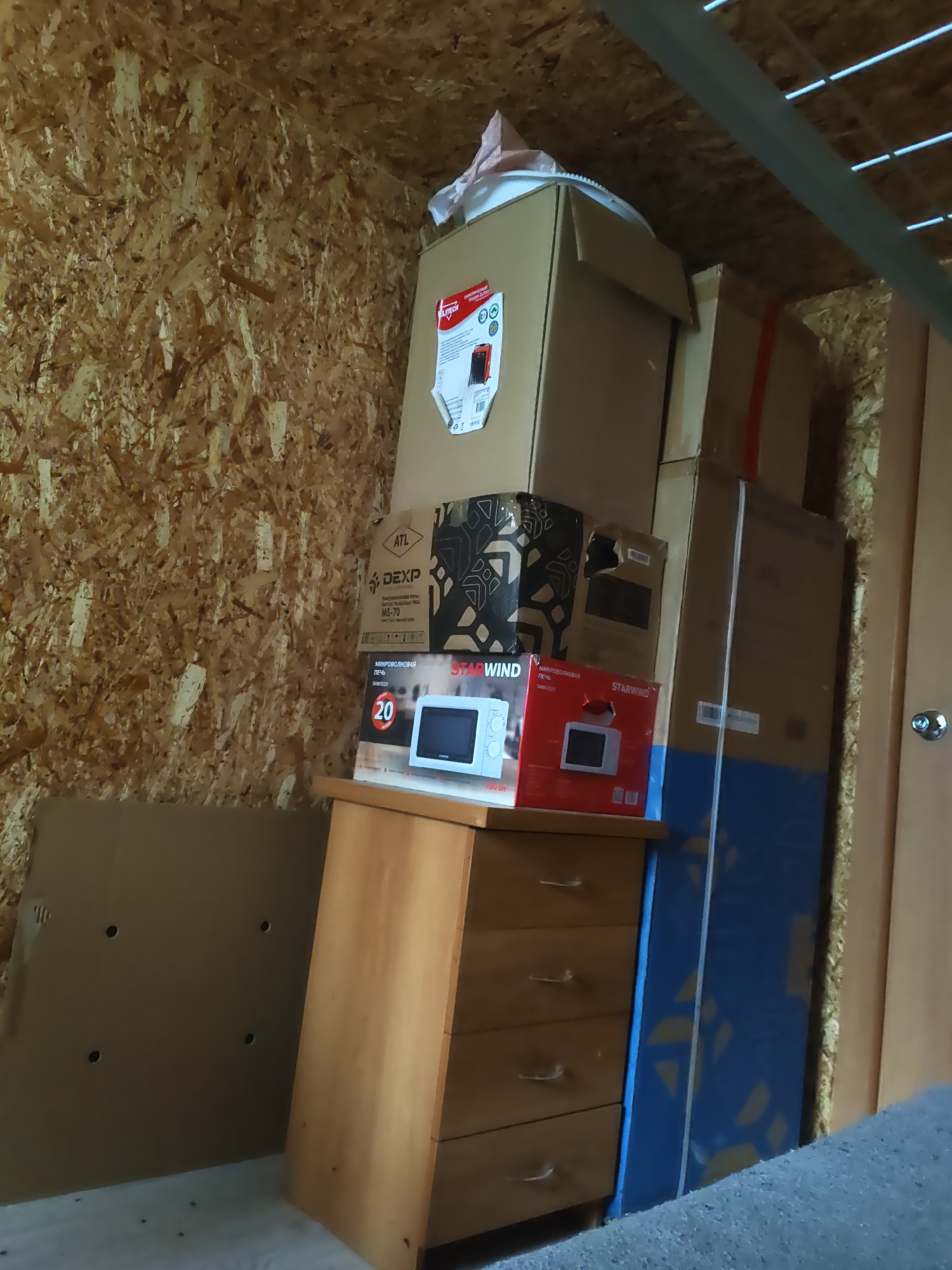 The image depicts a storage area, possibly a shed or attic, with walls constructed of oriented strand board (OSB). A light-colored ceiling with metal beams is visible at the top. The scene is filled with stacked cardboard boxes of various sizes. On the left, a large flat cardboard object leans against the OSB wall, with visible holes. In the center, a wooden dresser with four drawers is placed in front of the stacked boxes. The top of the dresser holds two boxes containing microwave ovens, one with a white and red design labeled "STARWIND" and another with a red design also labeled "STARWIND" and "МИКРОволновая ПЕЧЬ". Above these, another box with a red and white label is visible, along with a larger, predominantly tan cardboard box. Further to the right, a tall, narrow blue and yellow patterned object is leaning against the wall, adjacent to a closed wooden door with a silver knob on the right side of the frame. The floor appears to be concrete, covered in some areas with a grey, textured material. The overall impression is one of stored goods in a utilitarian space.