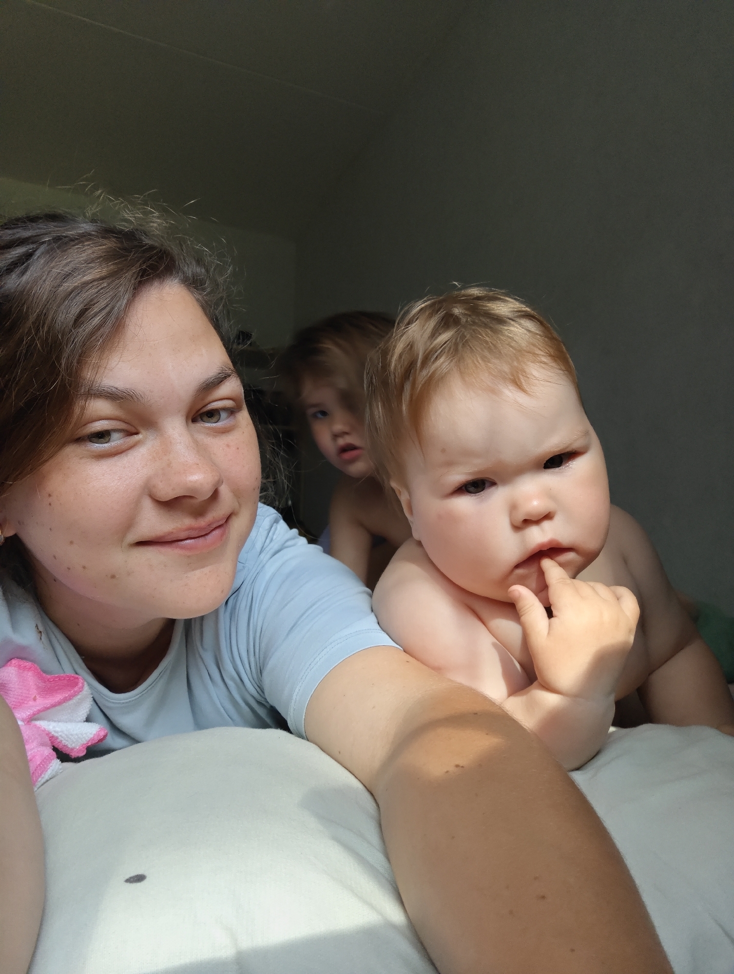The image captures a selfie of a woman lying on a white pillow. The woman is fair-skinned with brown hair pulled back, revealing her face with freckles. She is wearing a light blue shirt. Behind her, two young children are visible. The child closest to the camera has fair hair and is resting on their stomach, with one hand near their mouth. The second child behind them has lighter hair and a more obscured facial expression. The background appears to be a dimly lit room. There is a spot on the pillow. The image was taken indoors, presumably in a residential setting. The image was taken in the city of Chaikovsky, Russia.