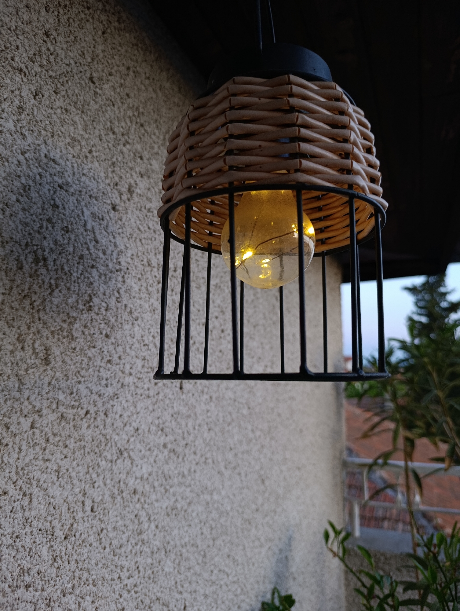 The image shows a decorative outdoor light fixture hanging near a building. The light has a woven basket-like shade over a metal cage, encasing a light bulb that is illuminated. The building's exterior wall has a rough, textured surface. The environment includes greenery, with hints of a balcony and other structures visible in the background. Without more specific information, it's impossible to confirm this location in Svilengrad, Bulgaria.
