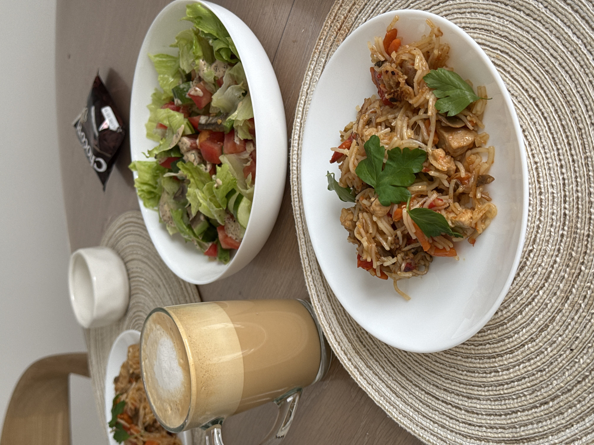 A close-up, eye-level shot showcases a meal and a beverage laid out on a wooden table in what appears to be a domestic setting in Gamovo, Russia. In the foreground, a white plate holds a serving of stir-fried noodles with chicken, carrots, and red peppers, garnished with fresh parsley. To the left, a clear glass mug contains a foamy coffee drink, likely a latte or cappuccino. Behind these, a white bowl overflows with a fresh salad composed of lettuce, tomatoes, and cucumbers, lightly dressed. A dark, individually wrapped snack, possibly chocolate, rests in the background. A small, white ceramic dish sits to the left of the salad bowl. The table is covered with woven placemats under the plate and mug. The overall scene suggests a casual meal enjoyed indoors.