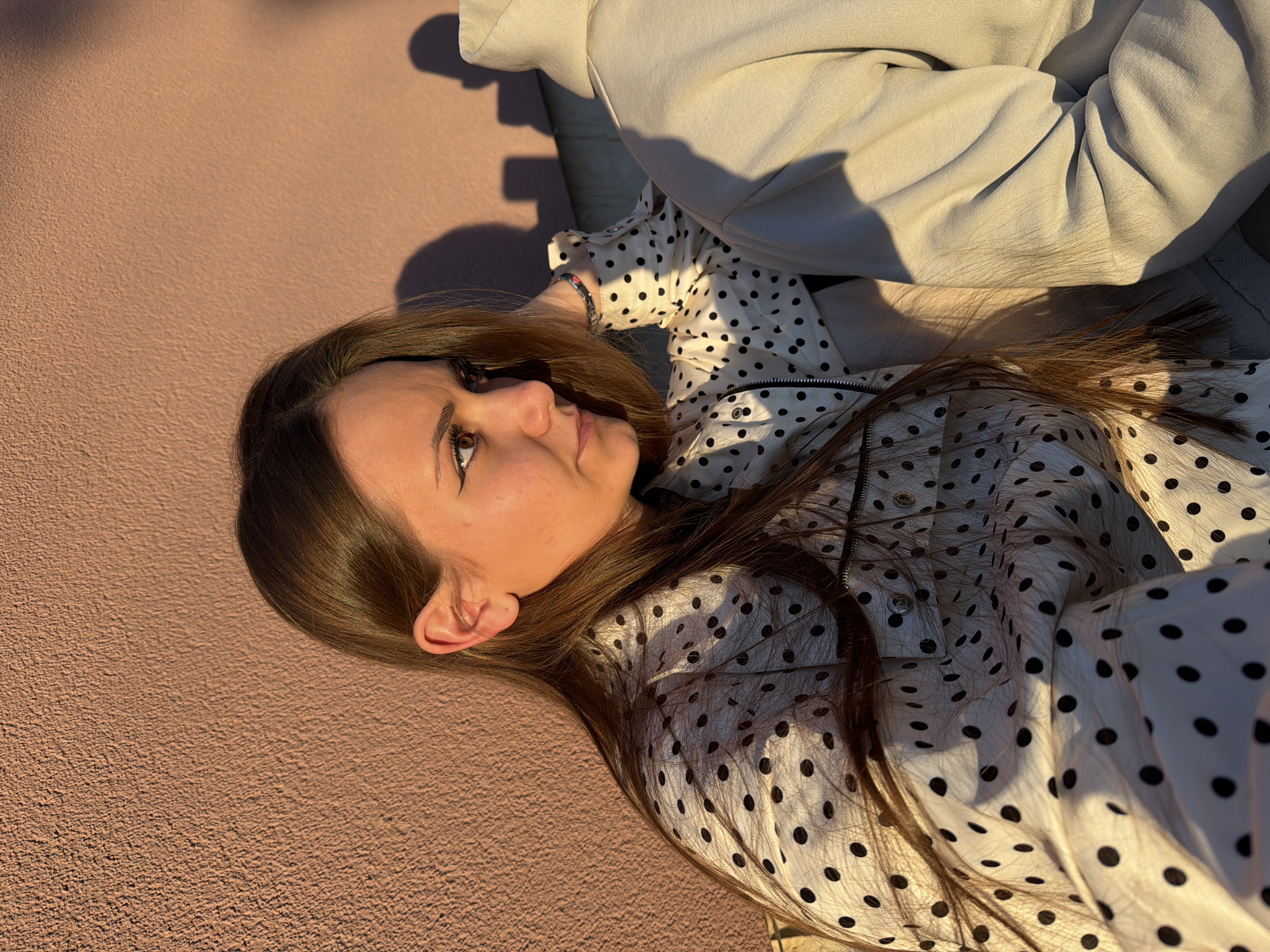 A woman with long brown hair is lying on a textured, possibly concrete, surface. She is wearing a white shirt with black polka dots. Part of a grey hooded sweatshirt is also visible in the upper right corner of the image. The lighting suggests it is daytime, possibly late afternoon. The location is not specified and cannot be confirmed as Buhovo, Bulgaria.