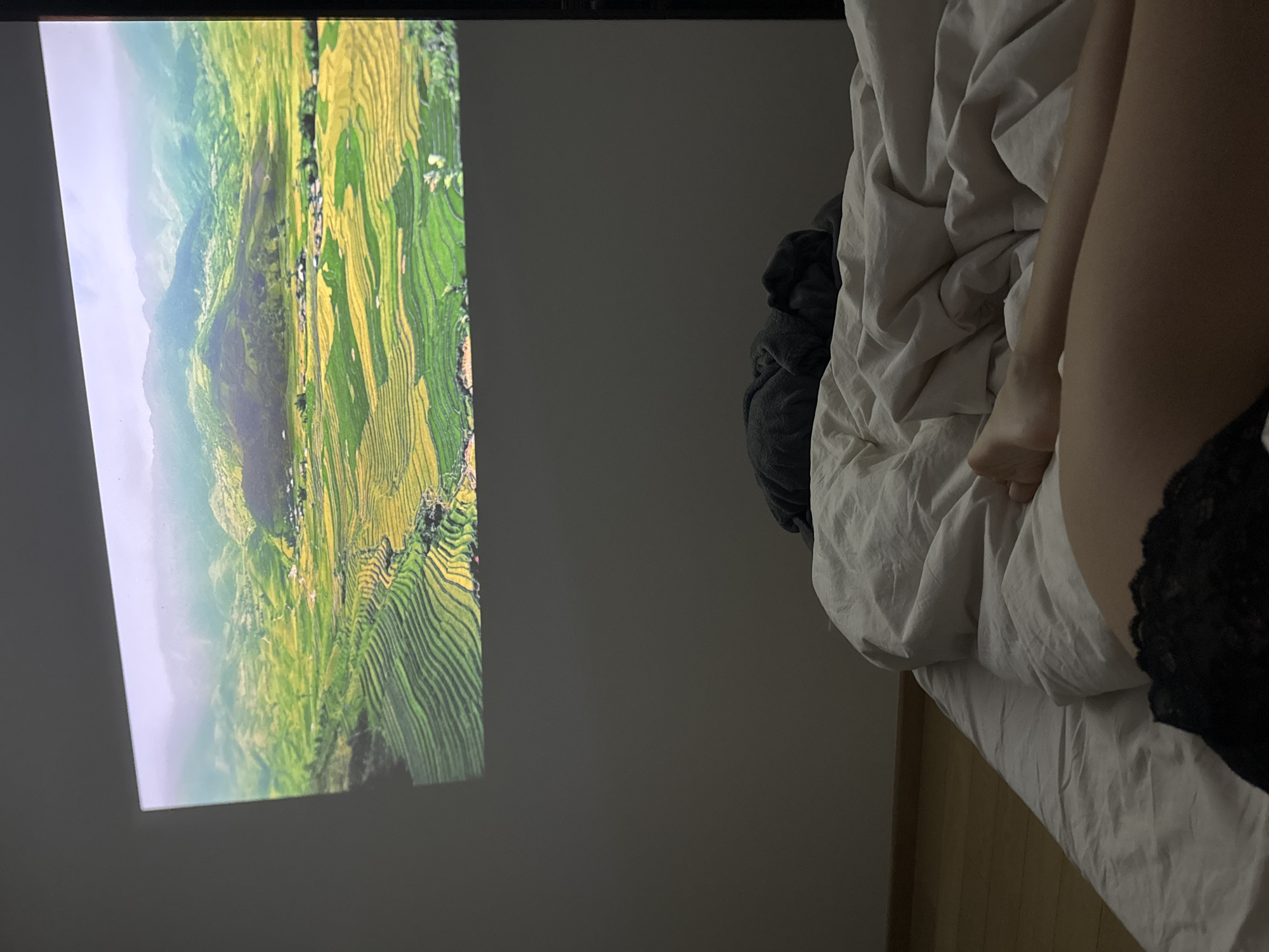 The image shows a view of a room with a nature scene projected onto the wall. The scene is composed of verdant fields and undulating hills, giving the impression of rice terraces, bathed in soft light. To the right of the projection is a bed with crumpled white linens. On the bed, a portion of a person's leg and thigh is visible, partially covered by black lace lingerie.
The image does not contain information allowing to determine the location. Therefore, it cannot be confidently stated that this image was taken in Warsaw, Poland.