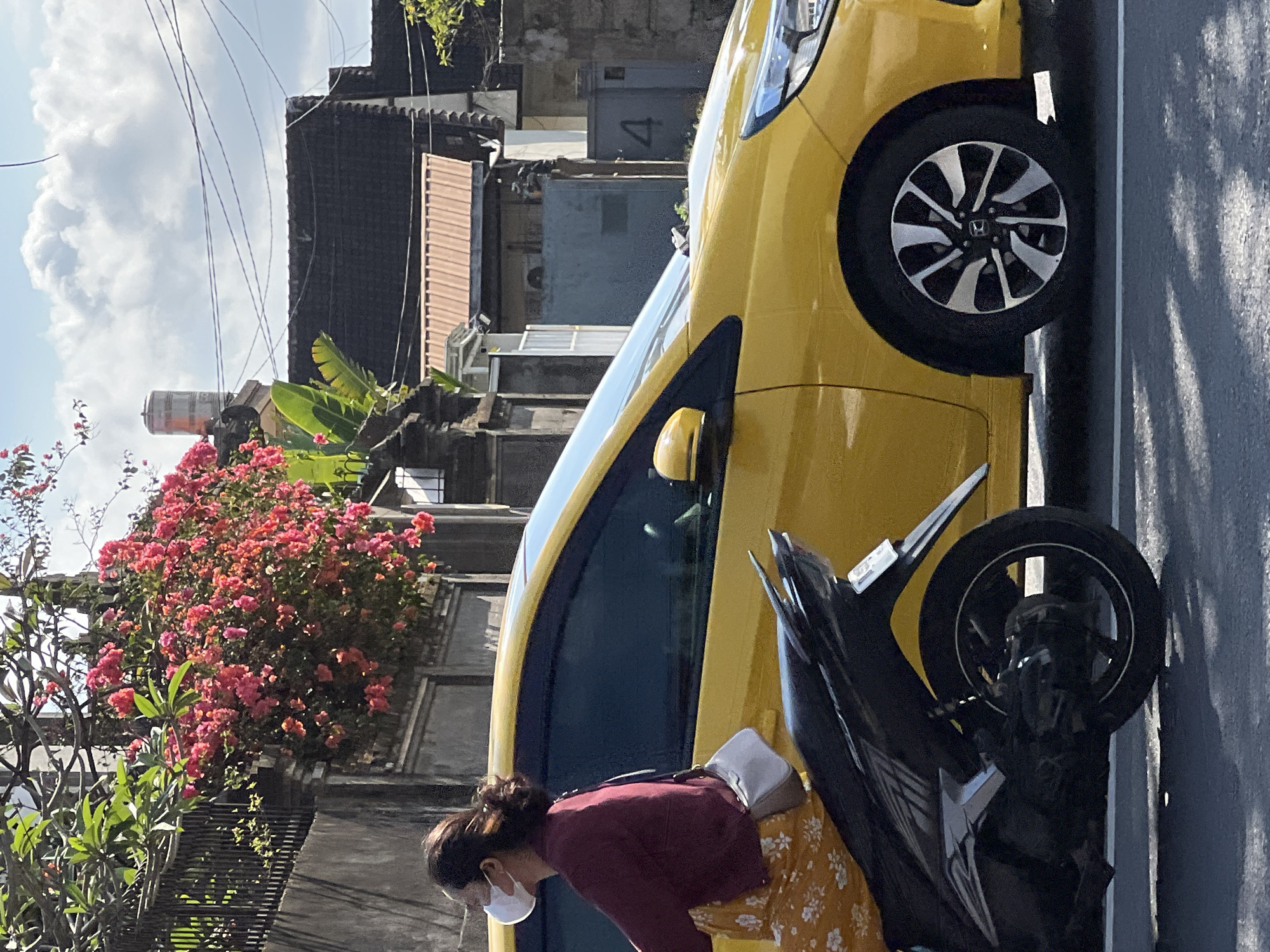 A person wearing a white face mask, a maroon long-sleeved shirt, and a yellow floral print skirt is riding a black motorcycle. The person is positioned on the left side of the frame, facing left. Behind the motorcycle is a yellow car, which occupies the majority of the right side of the image. The car is positioned on a paved road, with shadows cast on the road surface. In the background, there are buildings with tiled roofs, a gray wall, and lush green foliage with vibrant pink flowers cascading over a fence. The sky is partly cloudy with a bright blue sky visible. The scene appears to be in Denpasar, Indonesia, indicated by the architectural style, vegetation, and presence of a motorcycle commonly used as transportation in the region.