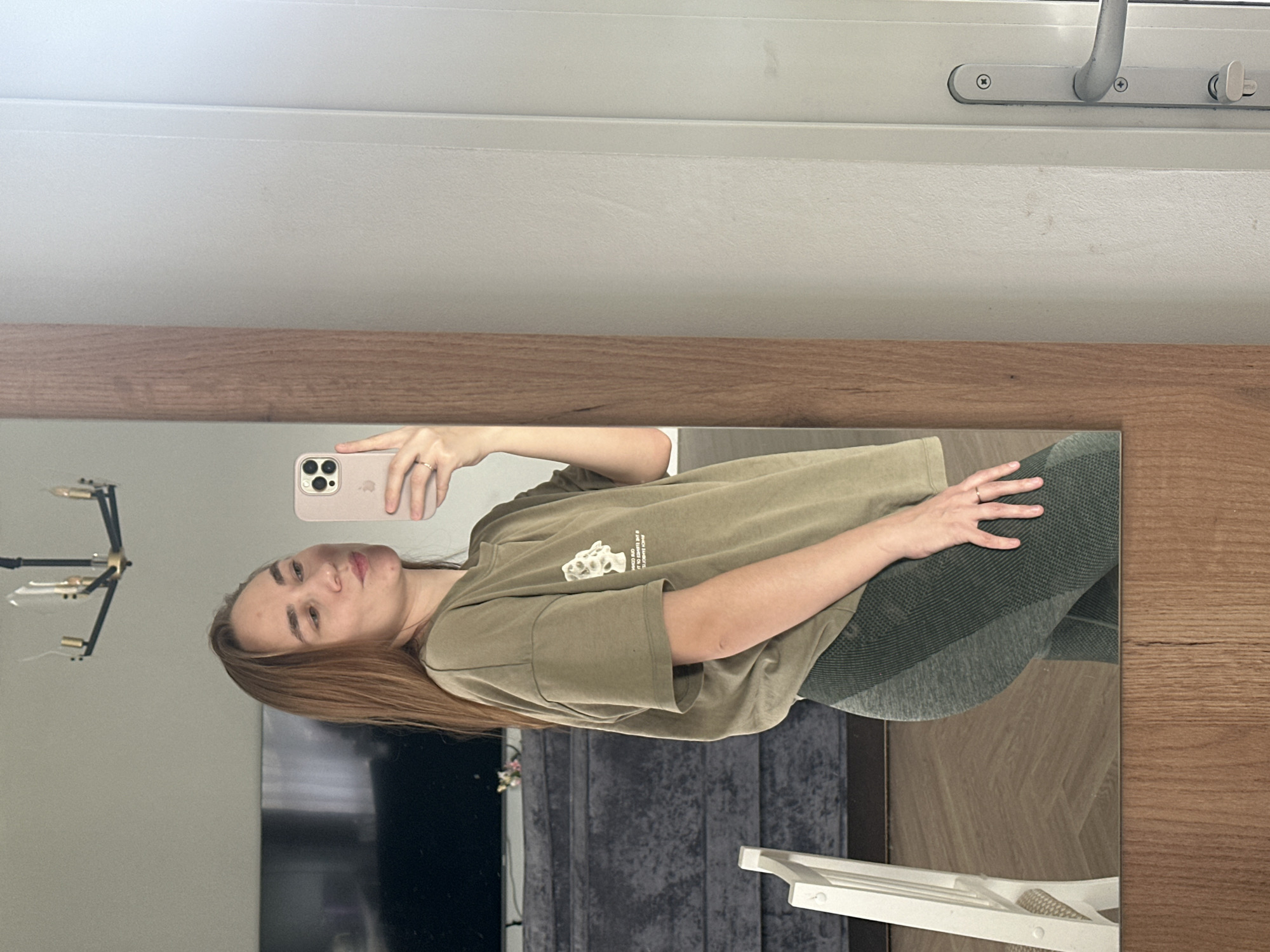 A young woman is captured in a mirror selfie, set indoors in Kharnavy, Russia. She stands before a large, rectangular mirror framed by a light brown wooden border. The woman has shoulder-length, straight reddish-brown hair and looks towards the smartphone she holds up with her right hand to take the photo. Her expression is neutral.
She is dressed in a casual outfit consisting of a loose-fitting, olive green or khaki short-sleeved t-shirt. White text and a small graphic are visible on the front of the t-shirt, with partially legible phrases including "IS THE ESSENCE OF LI..." and "ONE COMN...". She wears dark green or grey textured leggings on her lower body. A simple ring is visible on the ring finger of her left hand, which rests on her hip. The smartphone is light pink or beige, featuring a prominent camera module with multiple lenses.
Above the mirror, a white window frame with a metallic handle and latch is partially visible, suggesting the presence of natural light, indicating it is likely daytime. Reflections in the mirror reveal elements of the room behind her: a modern black chandelier or light fixture, a large dark rectangular object resembling a television screen, a dark grey textured surface that could be a wall or furniture, and a white, possibly woven, object on the floor. The floor reflected in the mirror appears to be light-colored wood. The overall setting suggests a casual, home environment.