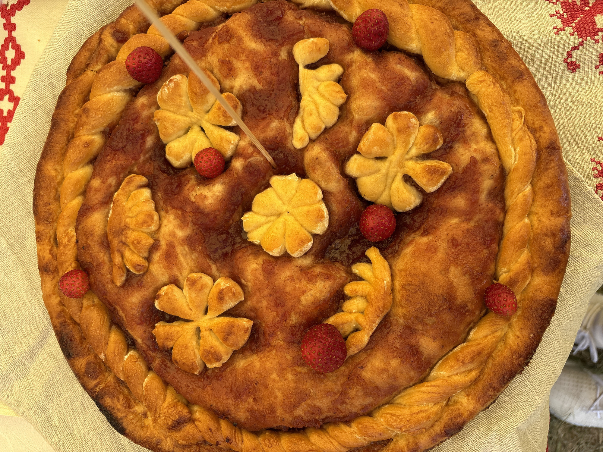 A golden-brown, round pie decorated with braided pastry edges, floral shapes cut from dough, and small, red wild strawberries sits on a beige, textured tablecloth. A skewer is inserted into the pie near the center. The pie appears to be filled with a dark, jam-like substance. In the lower left corner of the image, the tips of white sneakers are visible. The tablecloth has a red embroidered pattern along the right edge. The scene is lit from above, casting shadows on the pie's surface. The location is identified as Вяйнянскі сельскі Савет, Belarus.