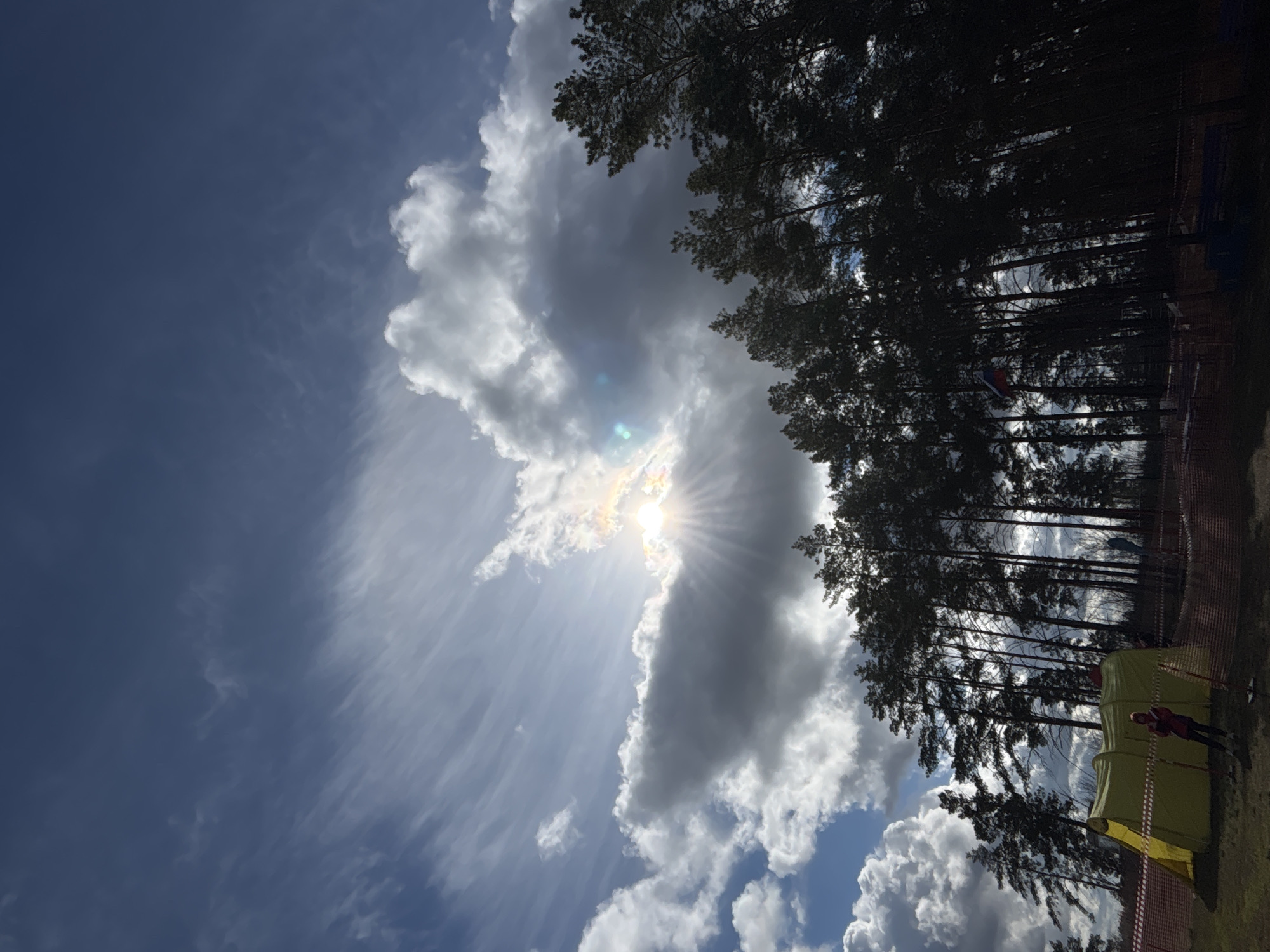 The image shows a scene in Orenburg, Russia. The sky is mostly blue with streaks of wispy clouds. The sun is shining brightly, partially obscured by a large, dark cloud formation that stretches diagonally across the frame. A dense line of tall trees is positioned on the right side of the image. A red-colored national flag can be seen flying atop of one of these trees. A couple of yellow and dark green tents are visible near the bottom right corner, next to a person wearing a red jacket standing on a grass field. A fence can be seen on the right side of the photo.