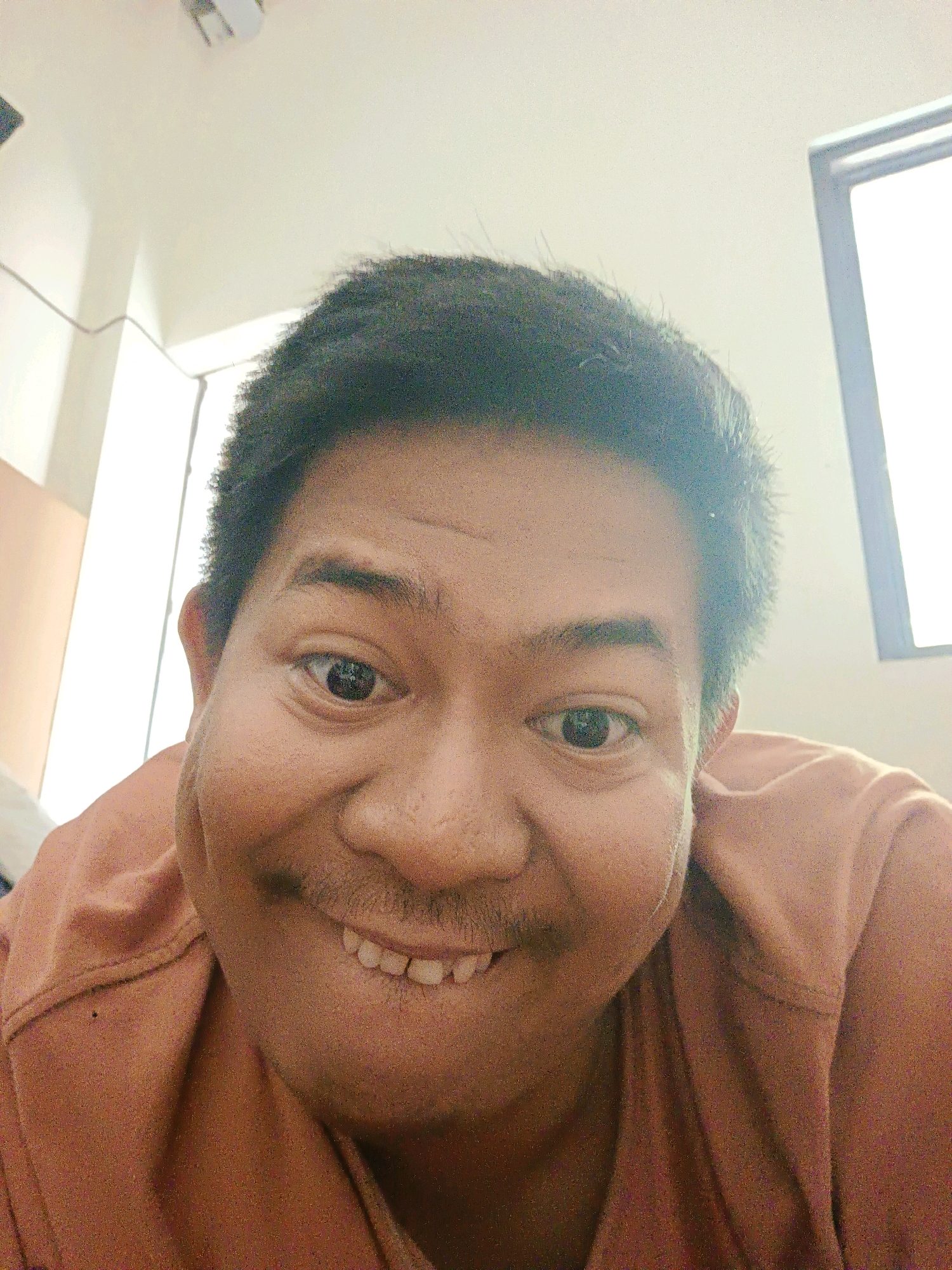 A low-angle, close-up, selfie-style shot shows a man's face from the nose up, with his head tilted forward and a wide, toothy grin. The man has dark, curly hair, dark eyes, and prominent dimples. He is wearing a rust-colored t-shirt. The background appears to be an indoor setting with white walls and a window with a white frame, suggesting it could be a room in Sragen, Indonesia, possibly an office or a residential space. The lighting is bright, coming from the top and right of the frame, creating a slightly washed-out effect.