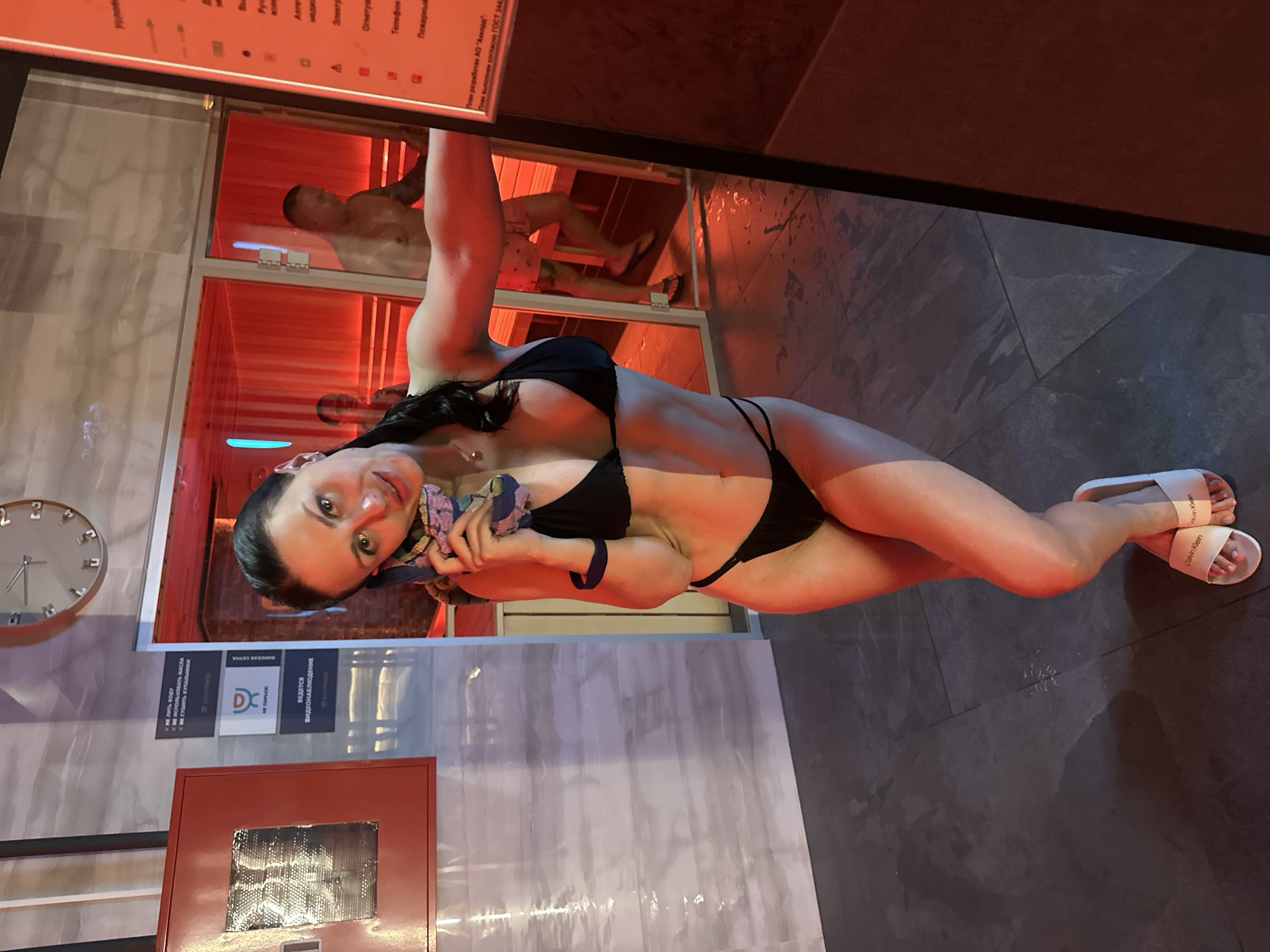 A woman in a black bikini stands in what appears to be a public sauna or spa facility in Novoe Devyatkino, Russia. She is holding a towel to her ear as if on a phone call. She is facing the camera with her left arm extended and her right hand holding the towel. Behind her, through a glass door, another person is visible inside the sauna. The environment is dimly lit with red lighting emanating from the sauna. The floor is dark tiled and appears wet in places. A clock is visible on the wall in the background. There are informational signs on the wall to the left of the woman, one of which has the logo "DEX FITNESS".